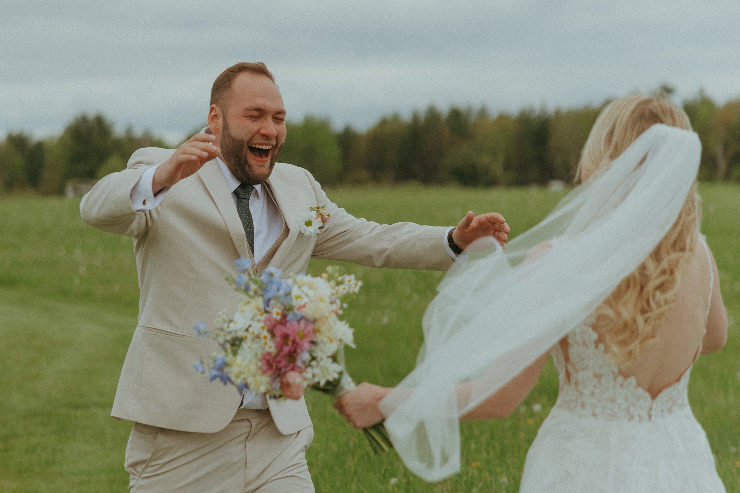 A man in a light-colored suit with a floral boutonniere laughing and reaching out to a woman in a white wedding dress and veil holding a bouquet in a green outdoor field.