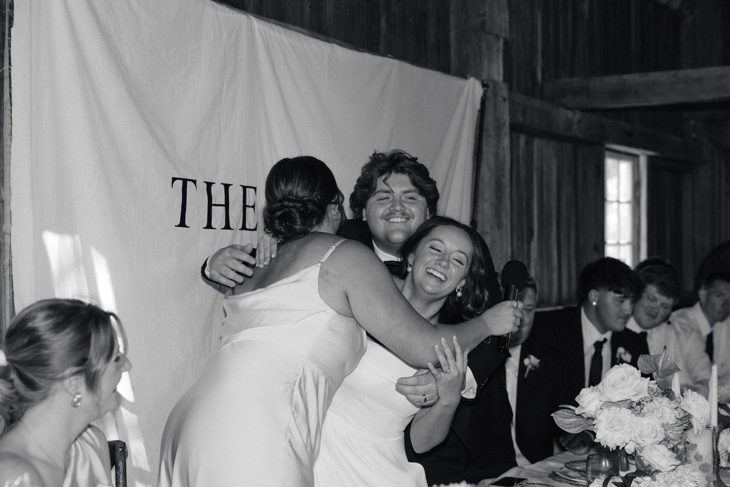 People hugging and smiling at wedding reception, with a bride holding a microphone, seated at a table with floral centerpiece, and friends in formal attire
