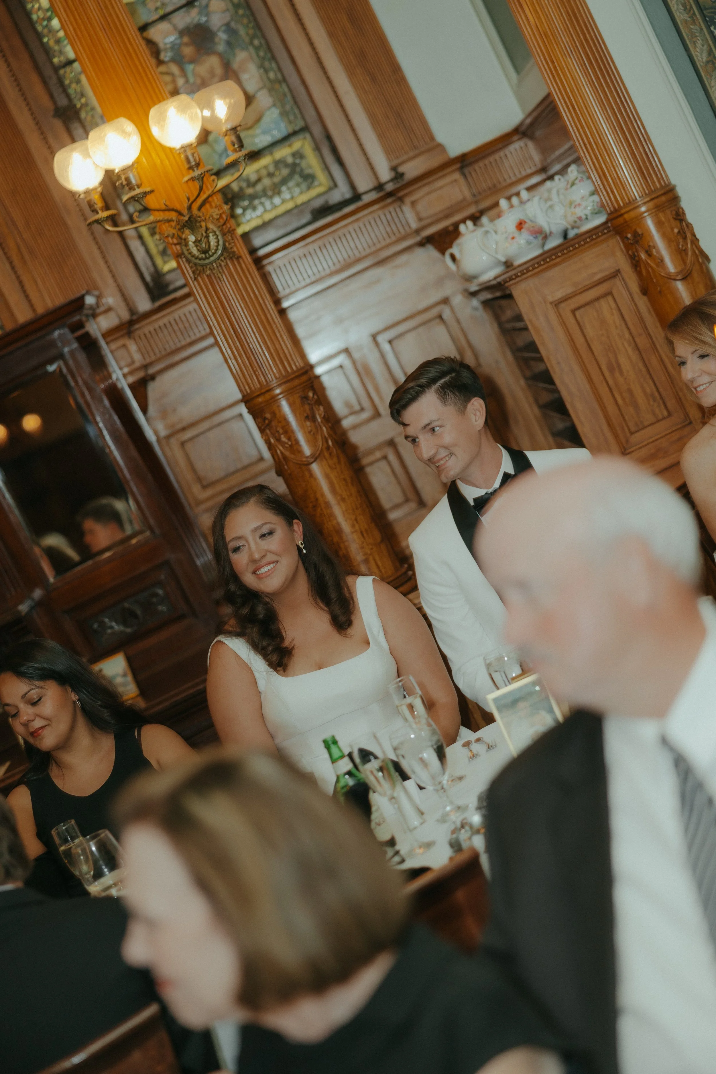 People in formal attire sitting at a table during a celebration, with wooden paneling and a chandelier in the background.