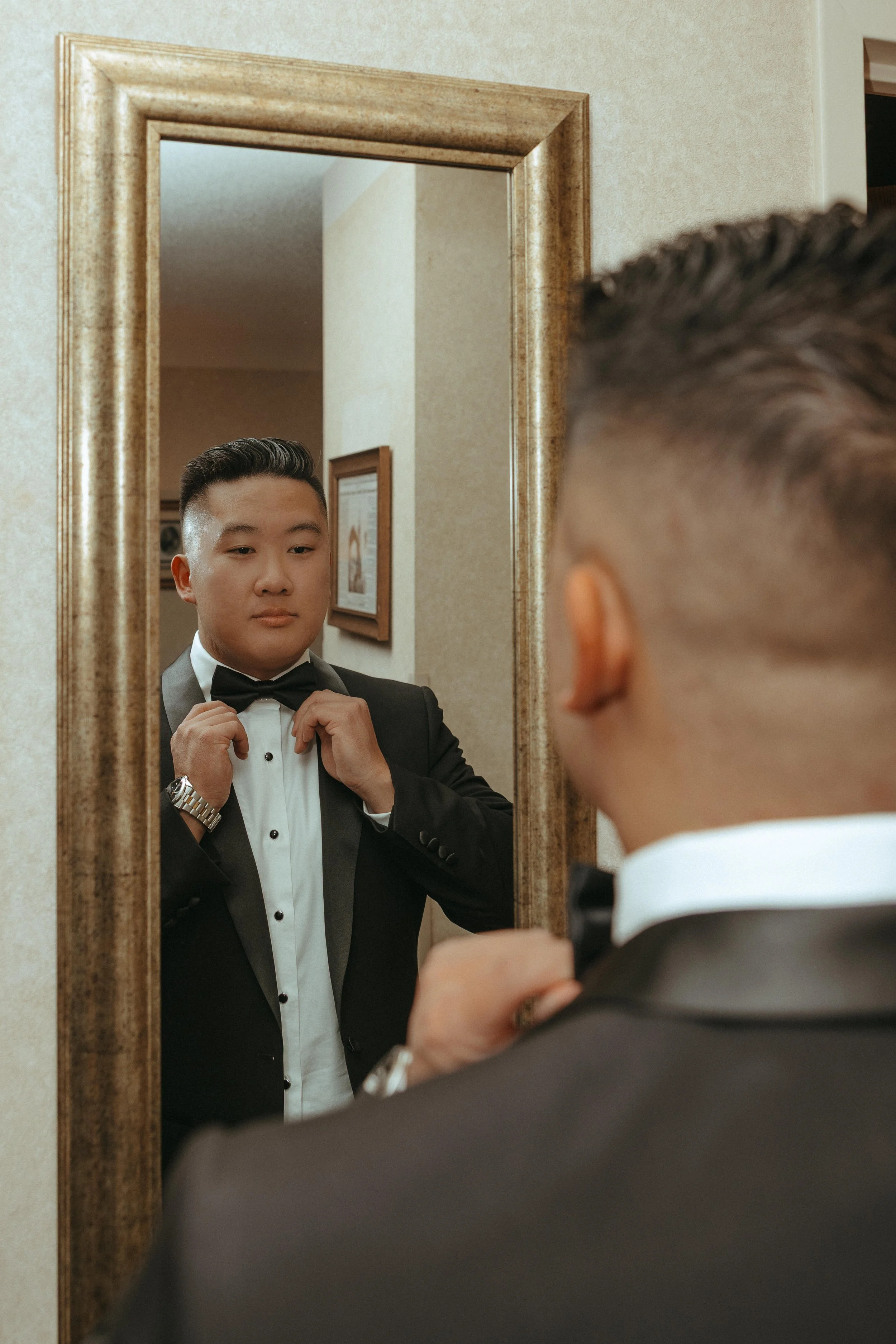 A young man in a formal tuxedo adjusts his bow tie in front of a mirror.