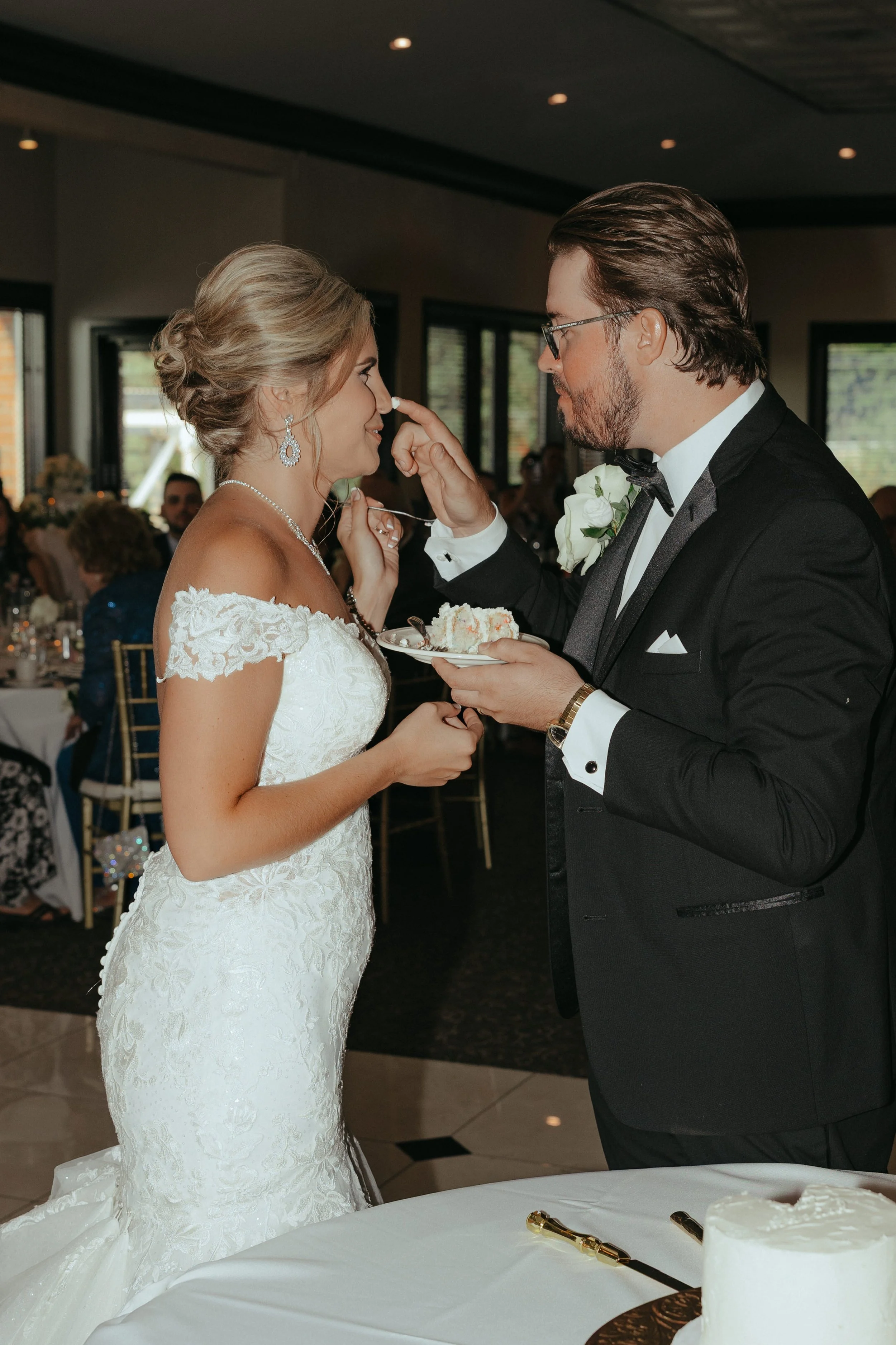 Bride and groom sharing wedding cake in a reception hall, with guests seated at decorated tables in the background.