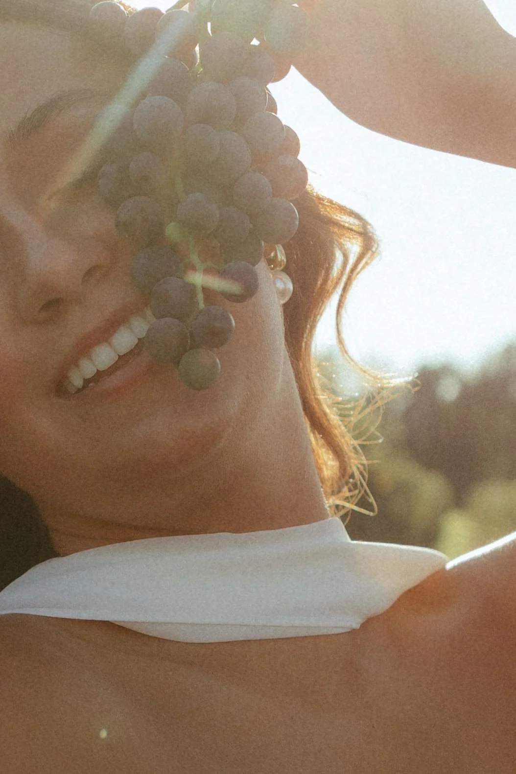 A woman with curly hair holding a bunch of grapes near her face, with sunlight creating a warm glow.