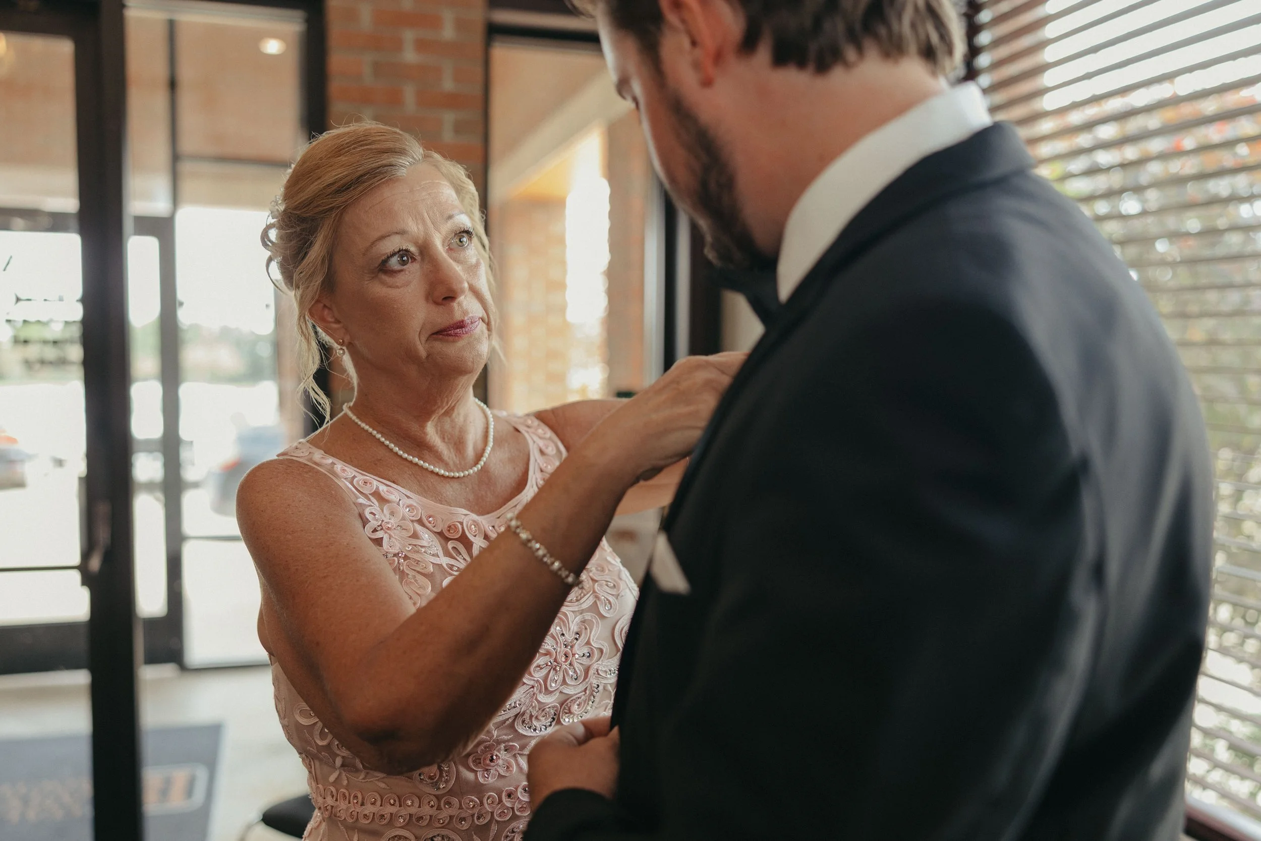 A woman in a pink lace dress and pearl necklace helping a man in a tuxedo adjust his jacket in a bright room with large windows.
