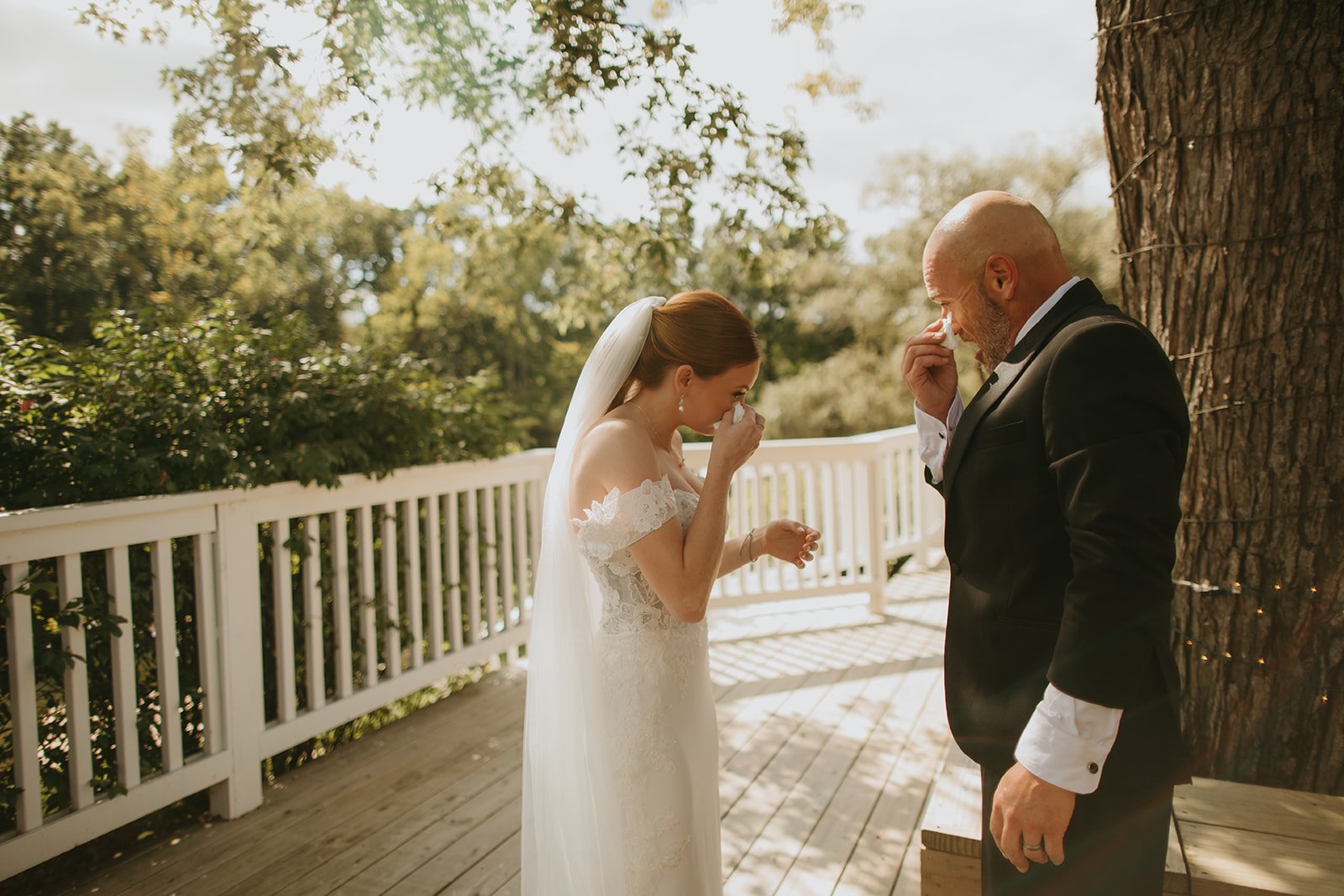 A bride and a groom crying tears of joy while holding tissues outdoors on a wooden porch with trees in the background.