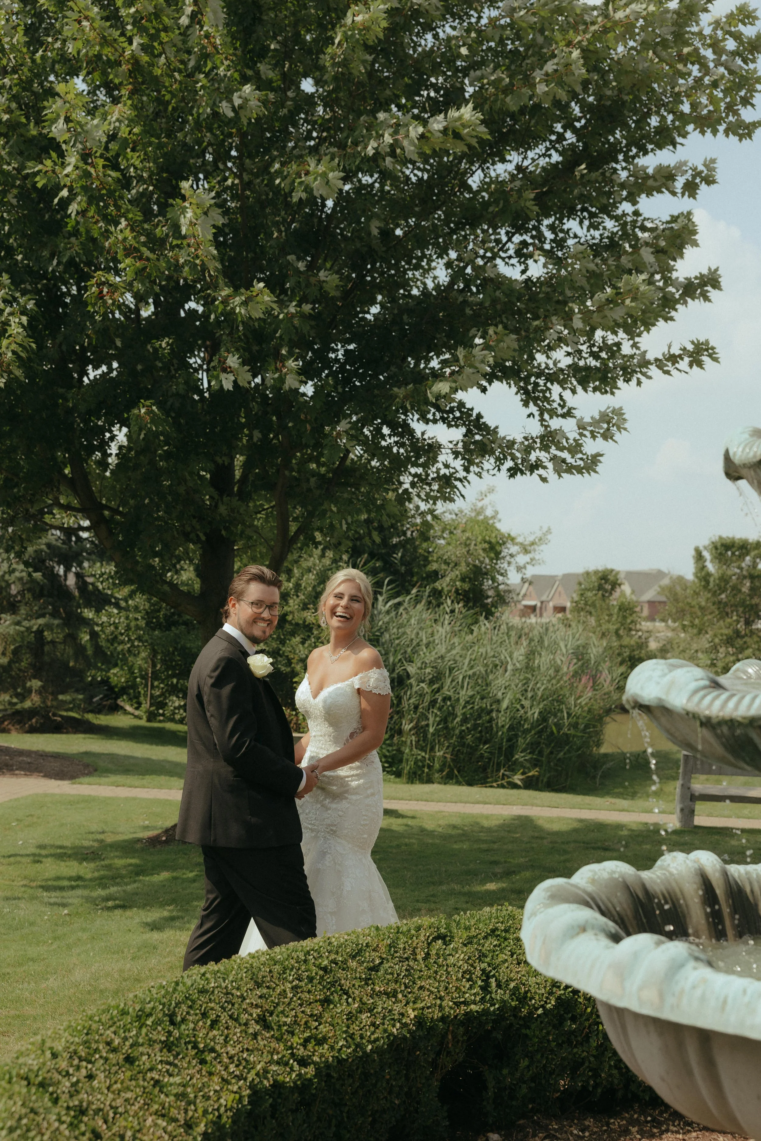 A bride and groom holding hands and smiling outdoors near a fountain and large tree, with a park and houses in the background.