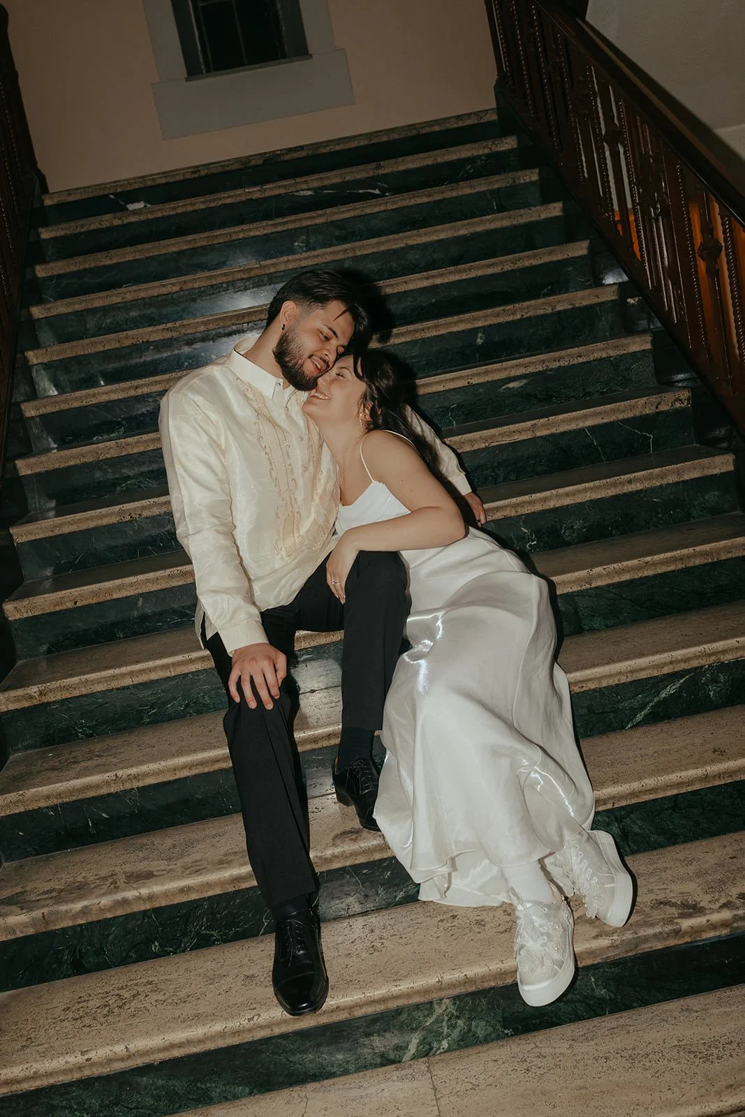 A couple in wedding attire lying on a staircase, smiling and affectionate, with the bride in a satin gown and sneakers, and the groom in a cream shirt and black pants.