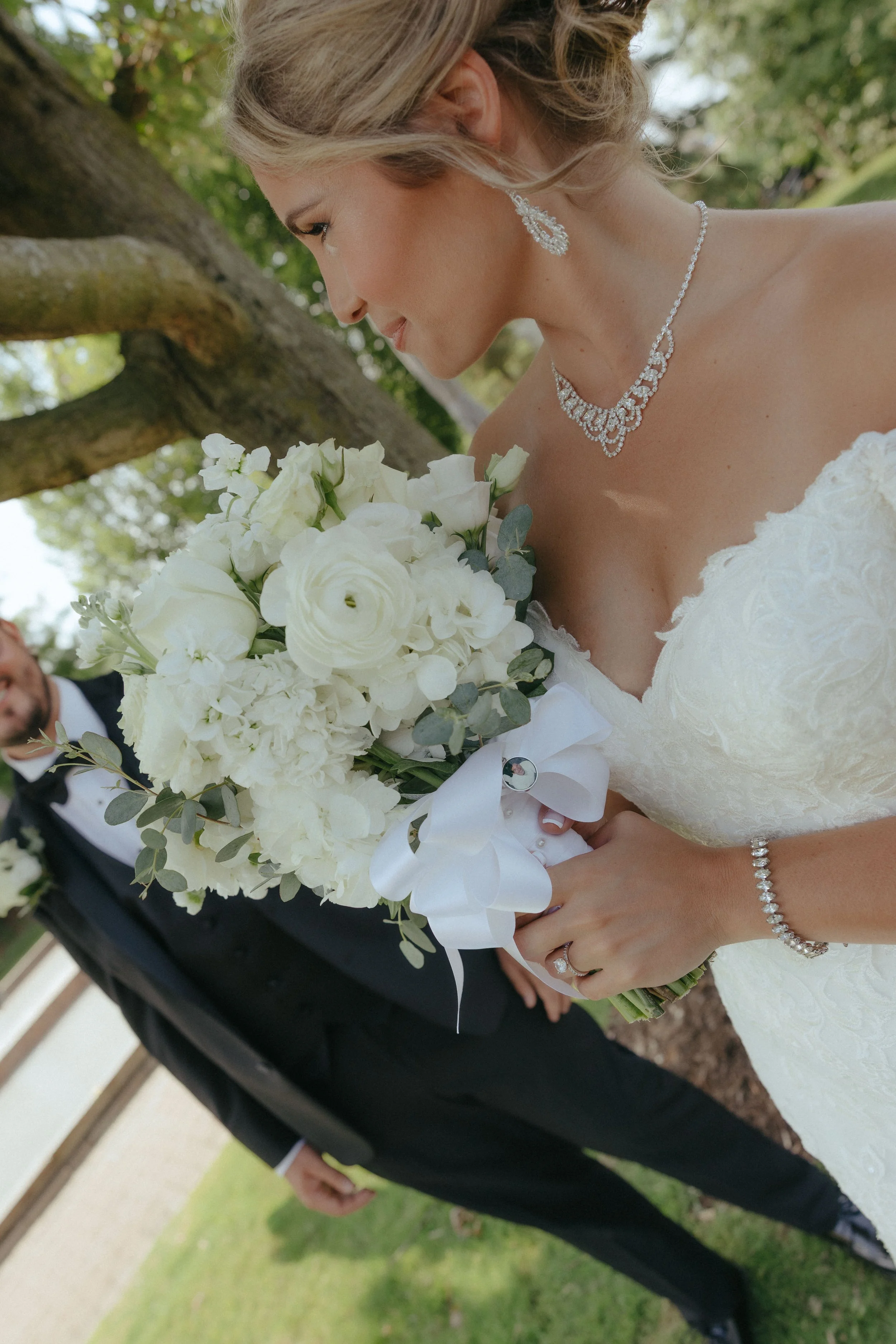 A bride in a white gown holding a bouquet of white flowers with green leaves, jewelry, and an updo hairstyle, standing outdoors near a tree.