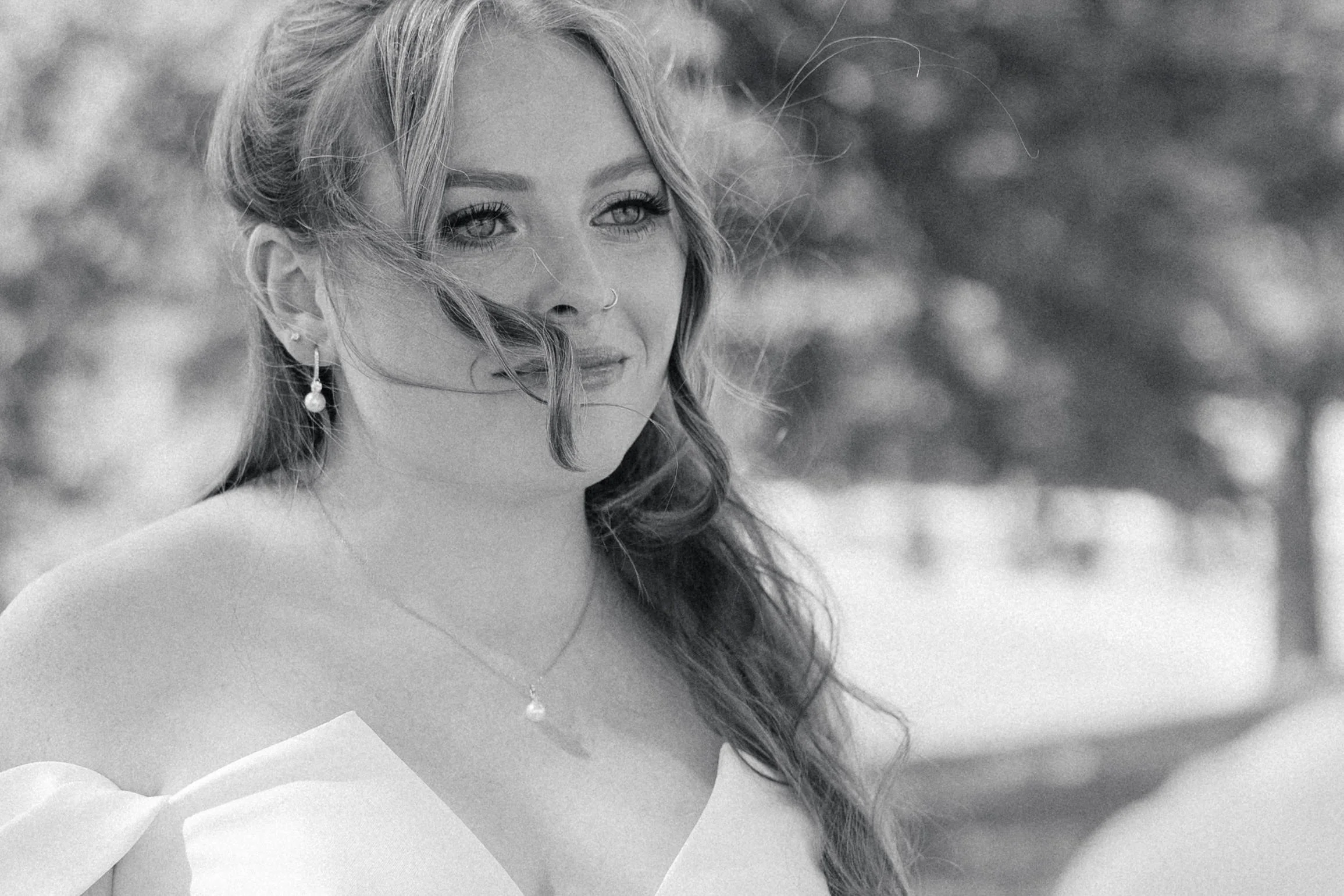 Black and white photo of a woman with long hair, wearing earrings and a necklace, outdoors with blurred trees in the background.