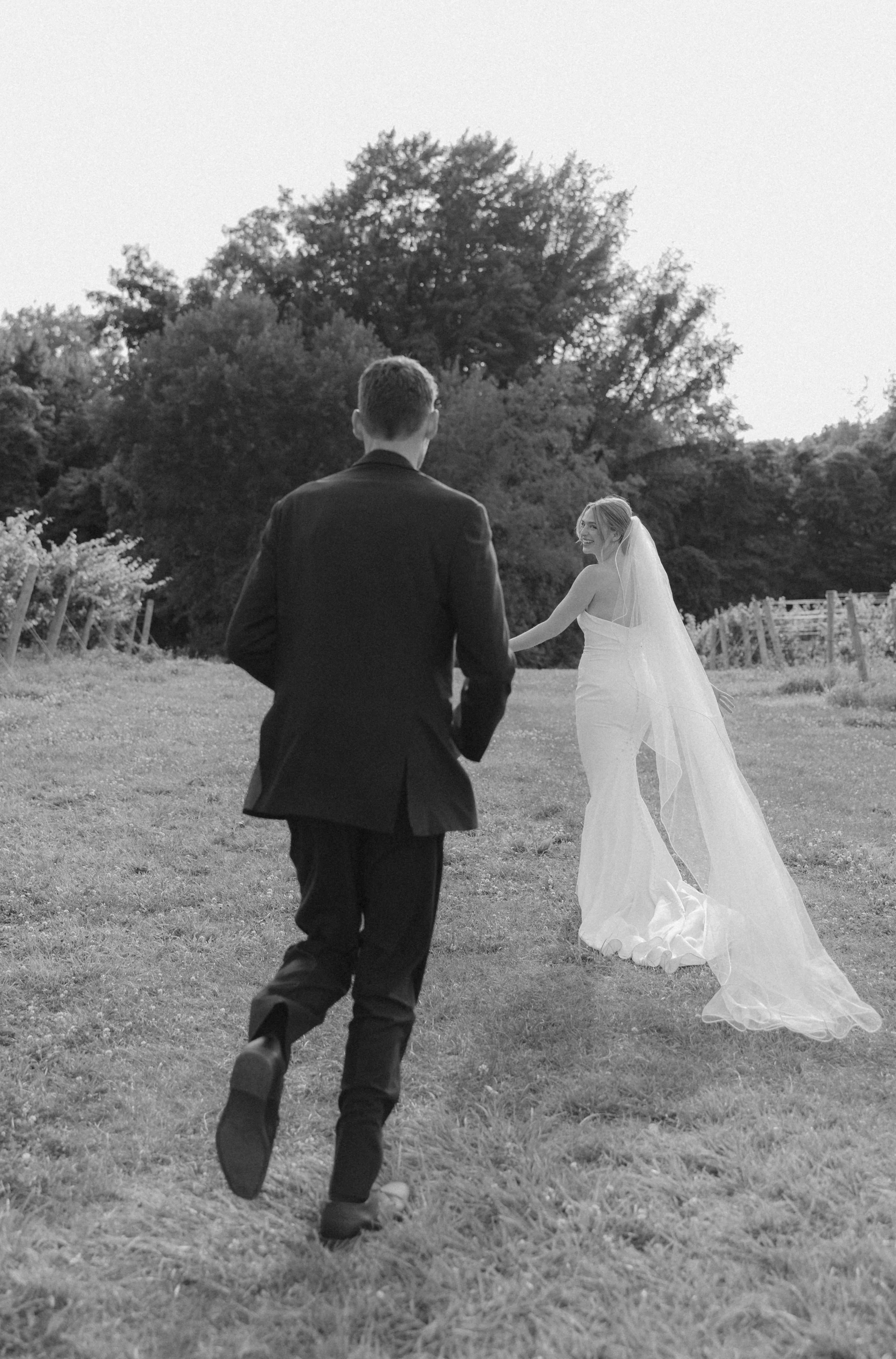 A black and white photo of a bride and groom outdoors. The groom is running towards the bride, who is smiling and holding his hand. The bride is wearing a wedding dress with a long train and veil, and the background features trees and a grassy field.