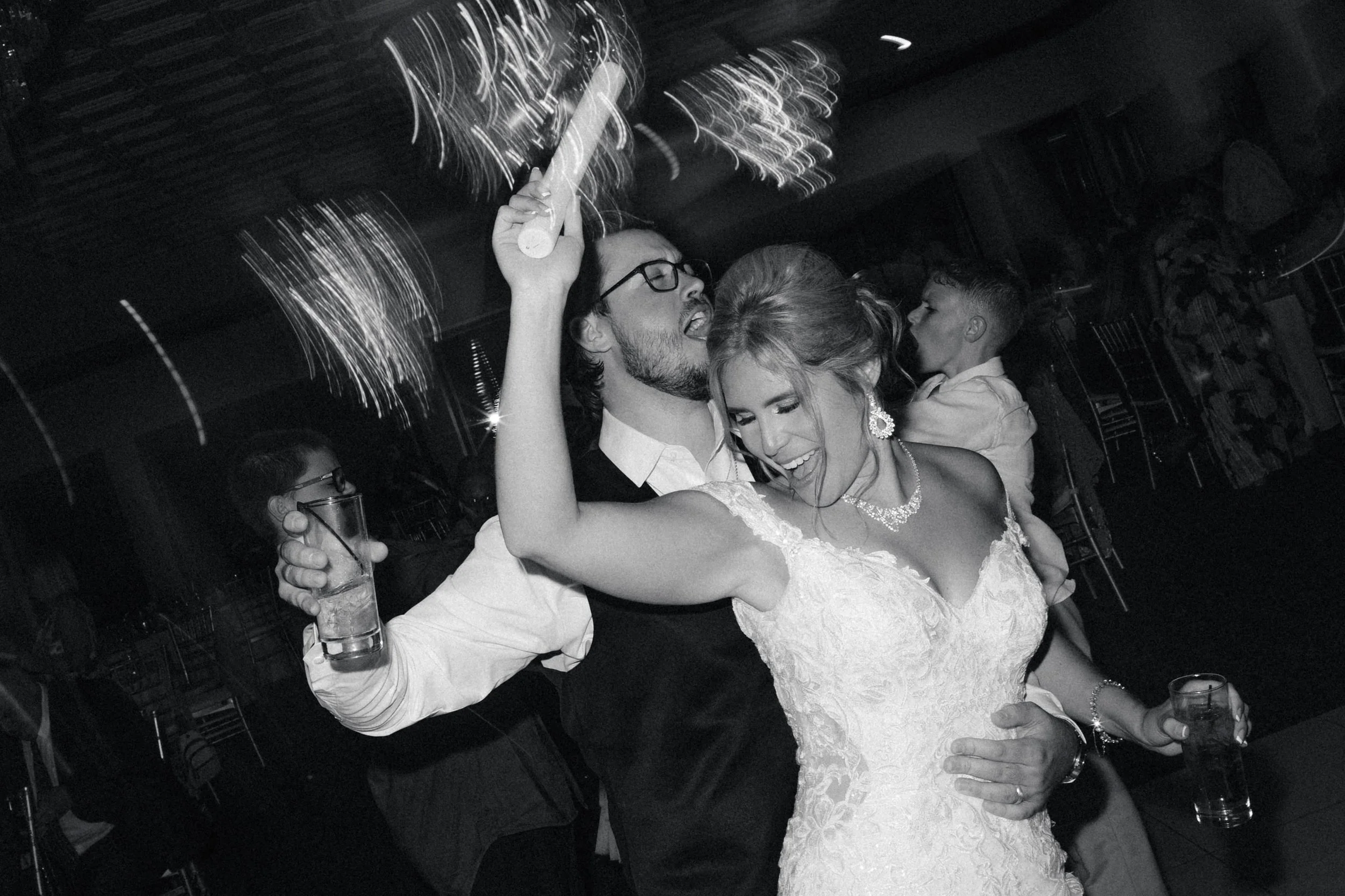 A wedding reception with a bride and groom dancing and celebrating together, surrounded by guests, in black and white.