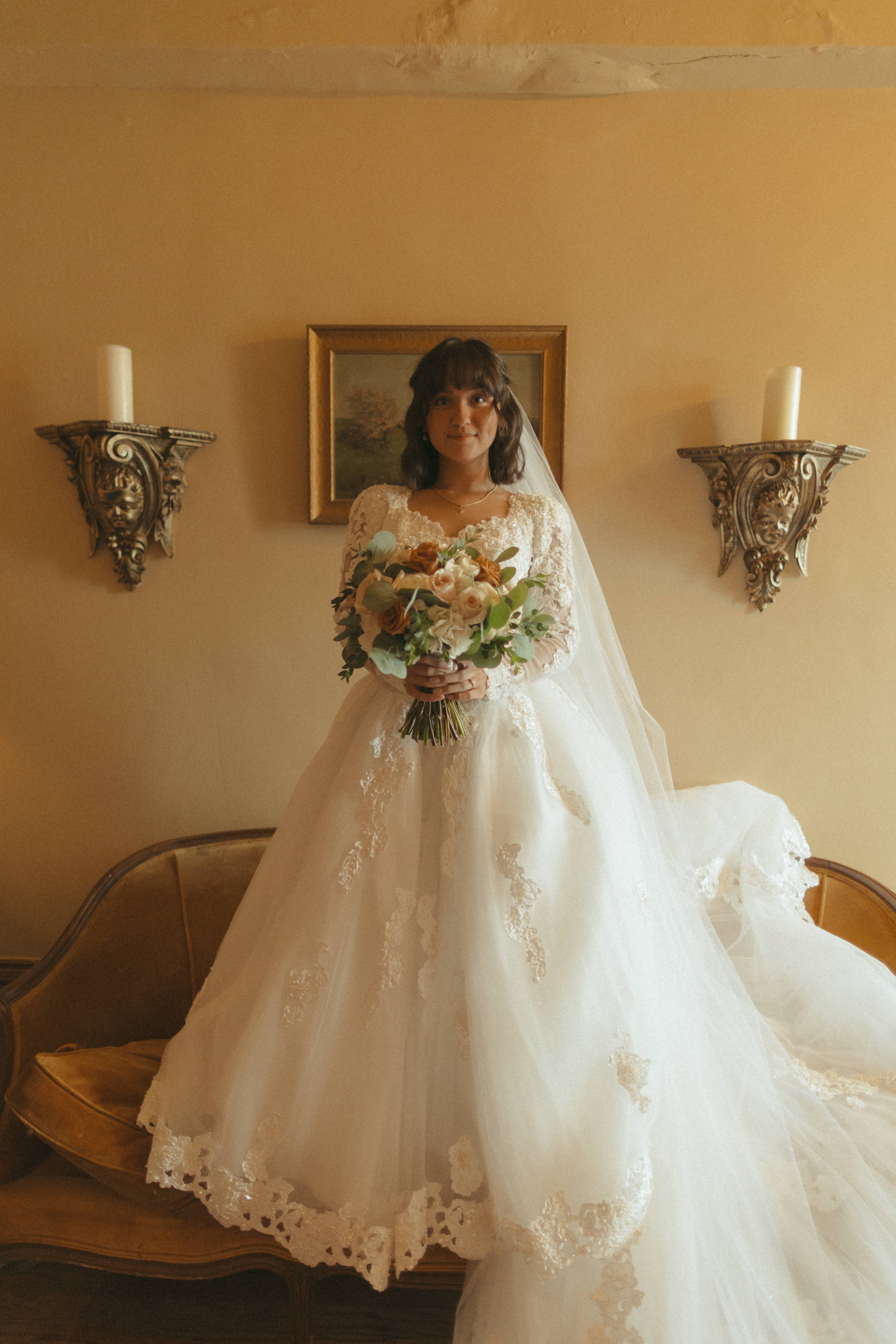 A bride in a white wedding gown holding a bouquet of flowers standing in a room with beige walls, two decorative wall sconces with candles, and a framed landscape painting.