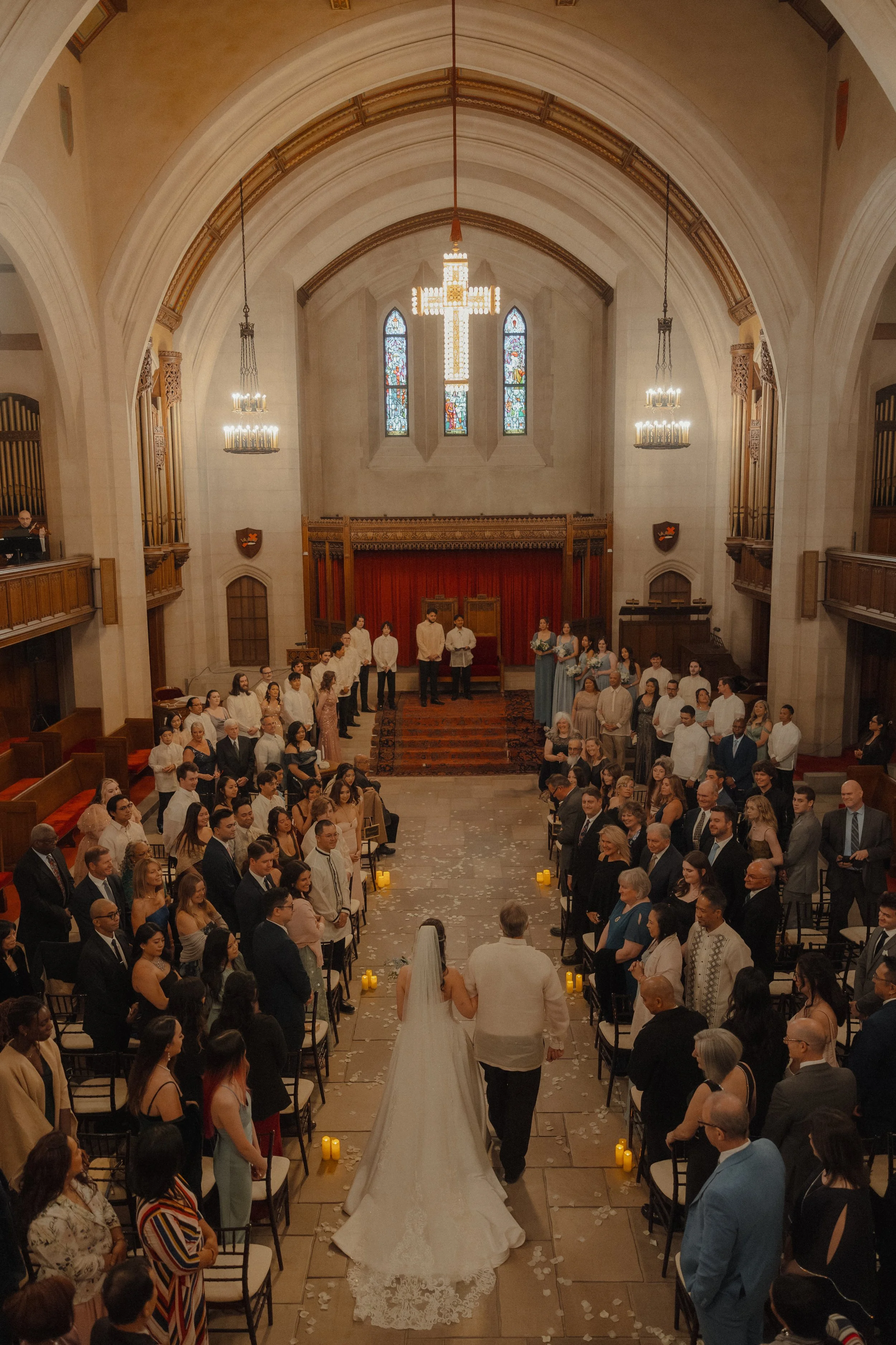 A couple walking down the aisle at their wedding ceremony inside a church, with guests seated and standing on either side, and a large chandelier hanging from the ceiling.