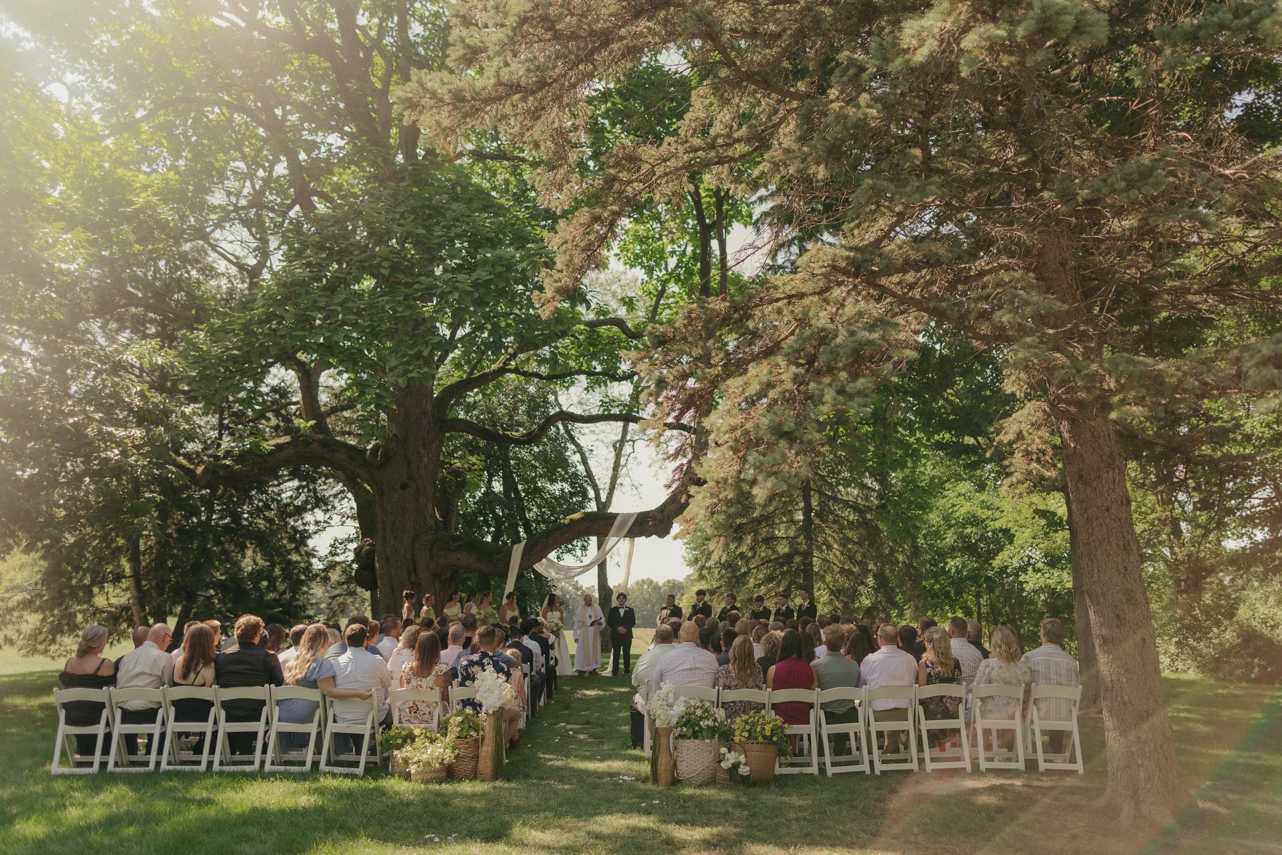 A large outdoor wedding ceremony under a big tree with many guests seated in white chairs, and the couple standing at the front with the officiant, surrounded by floral arrangements in baskets and pots.