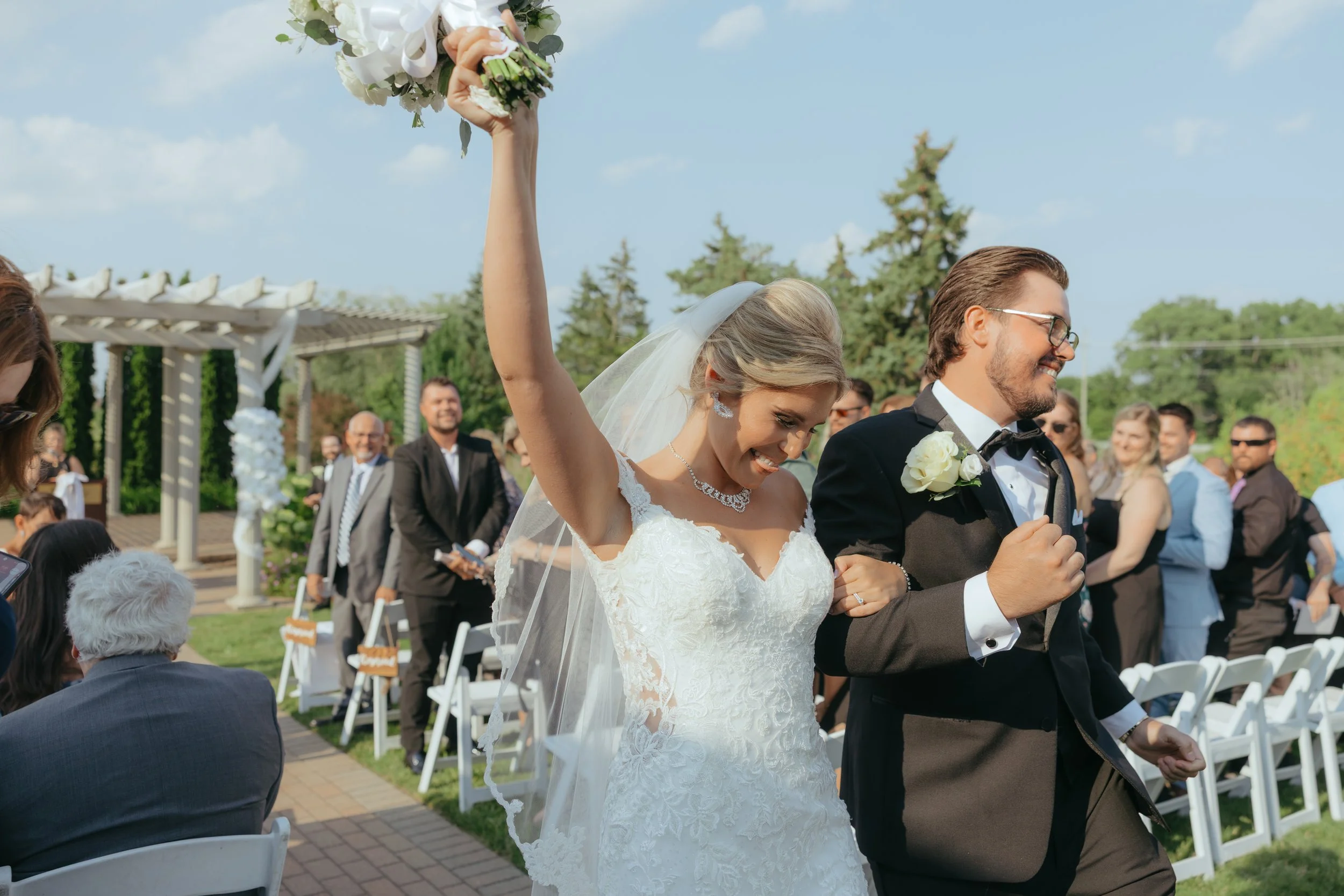 Bride and groom celebrating outdoors at their wedding, with the bride raising her bouquet and smiling, surrounded by guests.