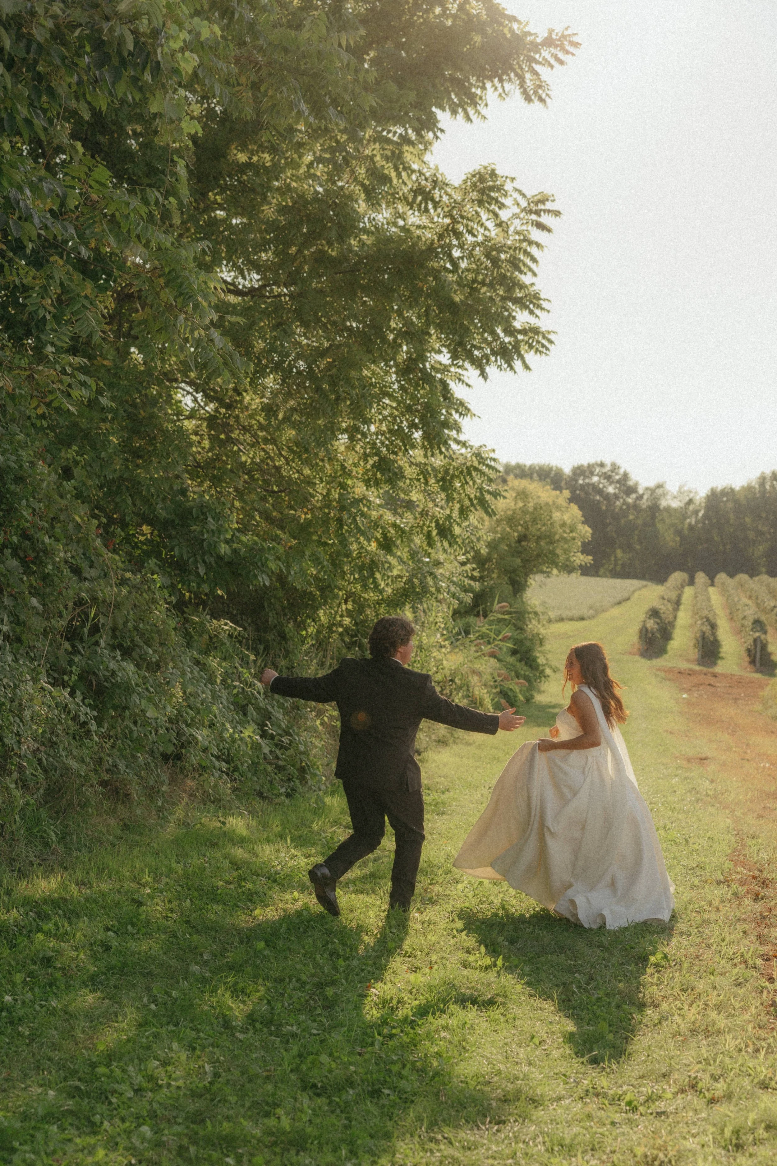 A bride in a white wedding gown is running towards a groom dressed in a black suit, on a grassy path through a green, wooded area with rows of farm crops in the background.