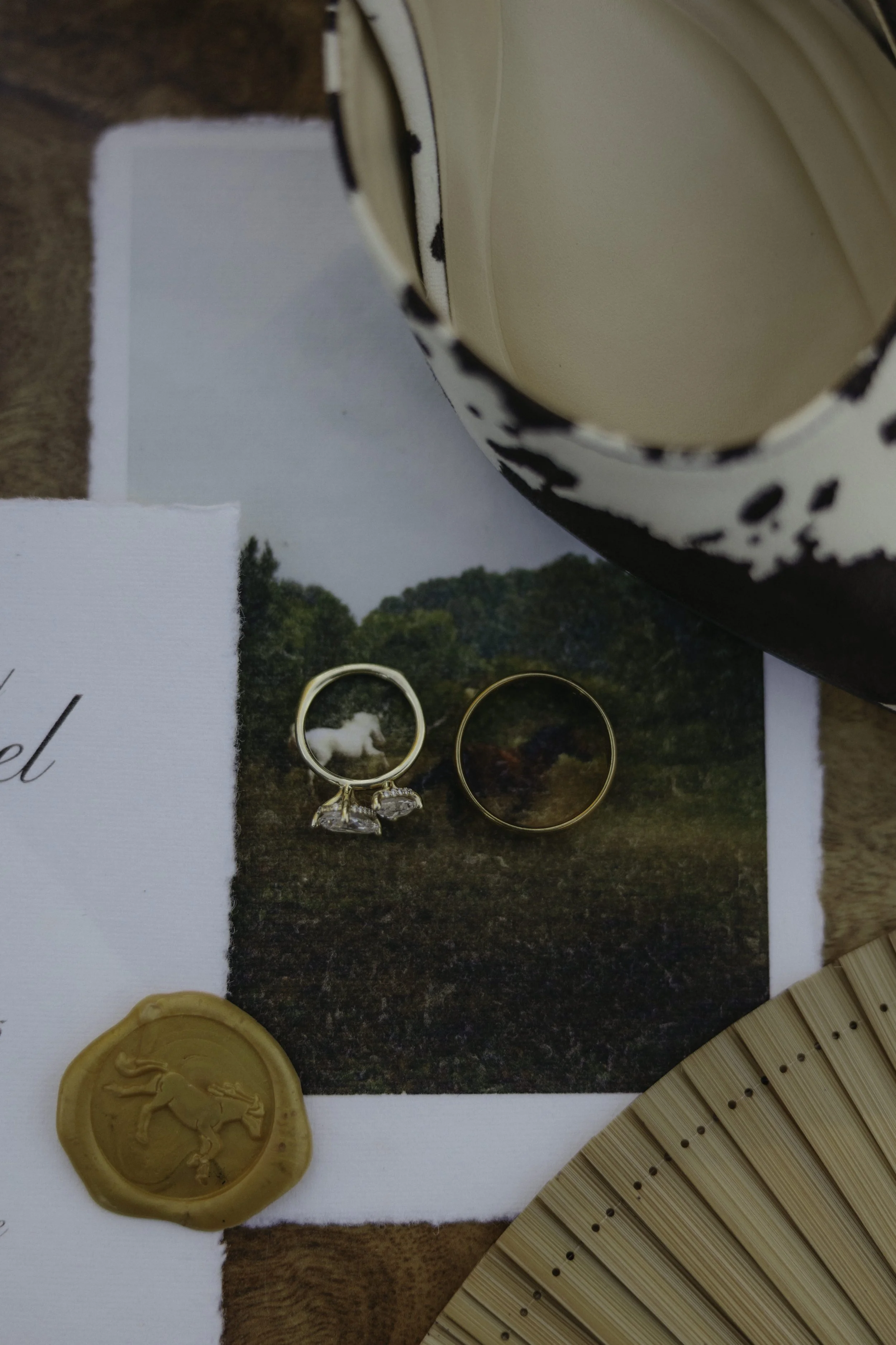 Close-up of a flat lay arrangement including a ceramic mug with Dalmatian print, a photo of a landscape with green trees, two rings (one gold with gemstones, one plain gold), a wax seal with a lion emblem, a section of a woven bamboo mat, and a handw