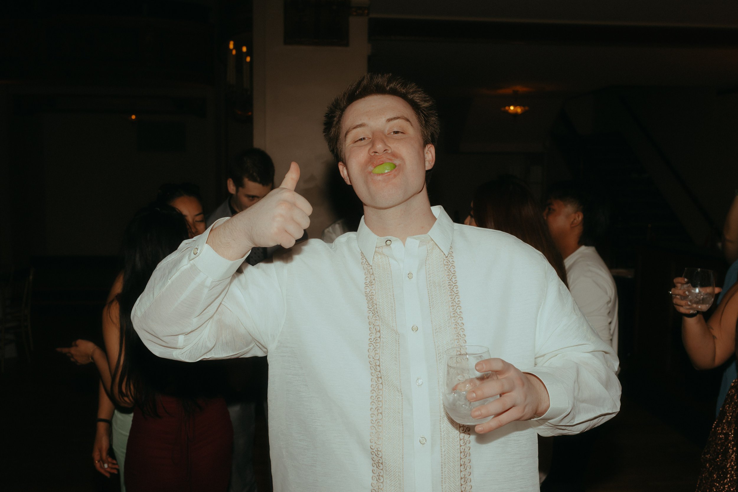 A young man in a white shirt holding a glass with a green lime in his mouth, giving a thumbs-up gesture at a social gathering or party.