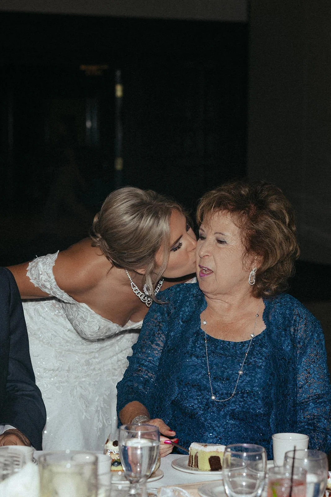 A woman in a white wedding dress kisses an older woman in a blue dress on the cheek at a celebration, likely a wedding reception, with a plate of cake and glasses on the table.