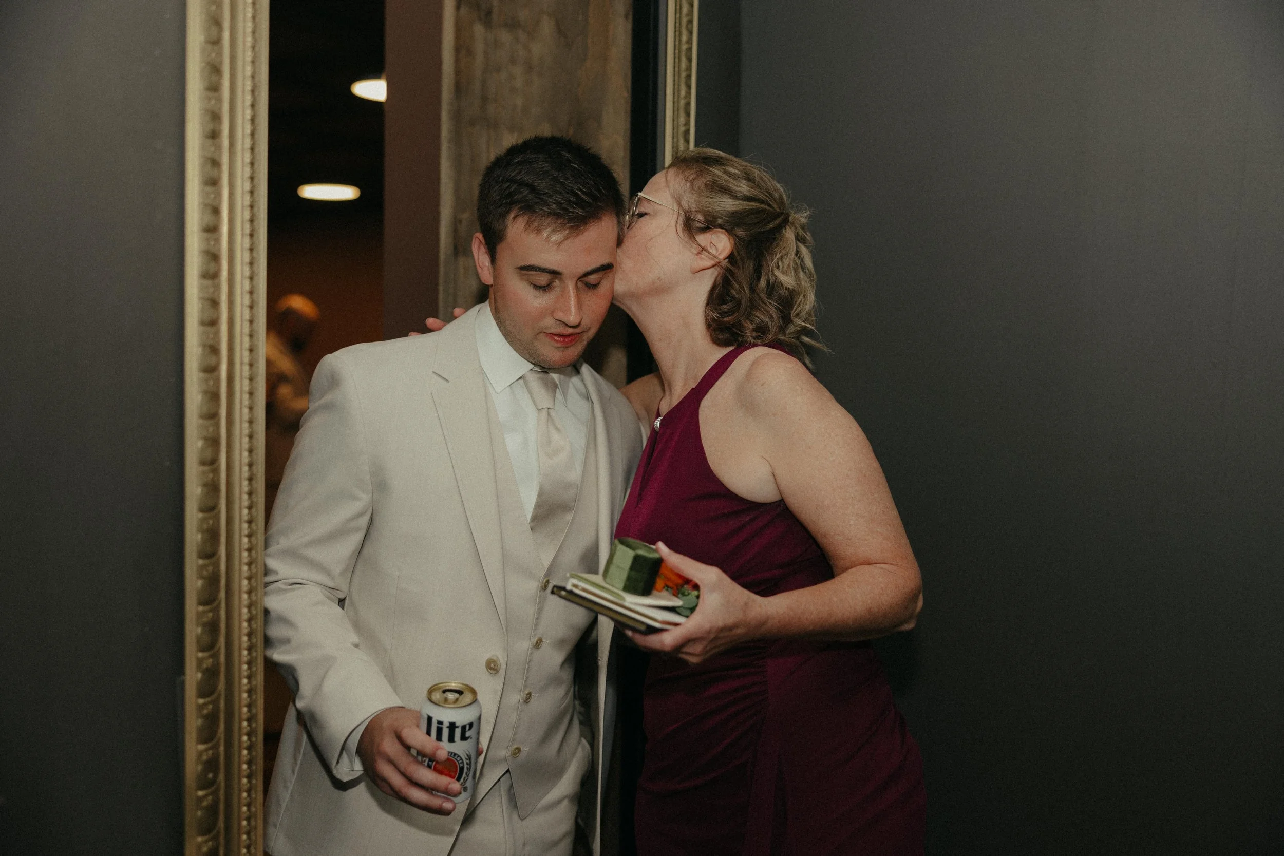 A woman in a burgundy dress kisses a man in a light-colored suit on the cheek in a dimly lit room, holding a small photo album and a can of Miller Lite beer.