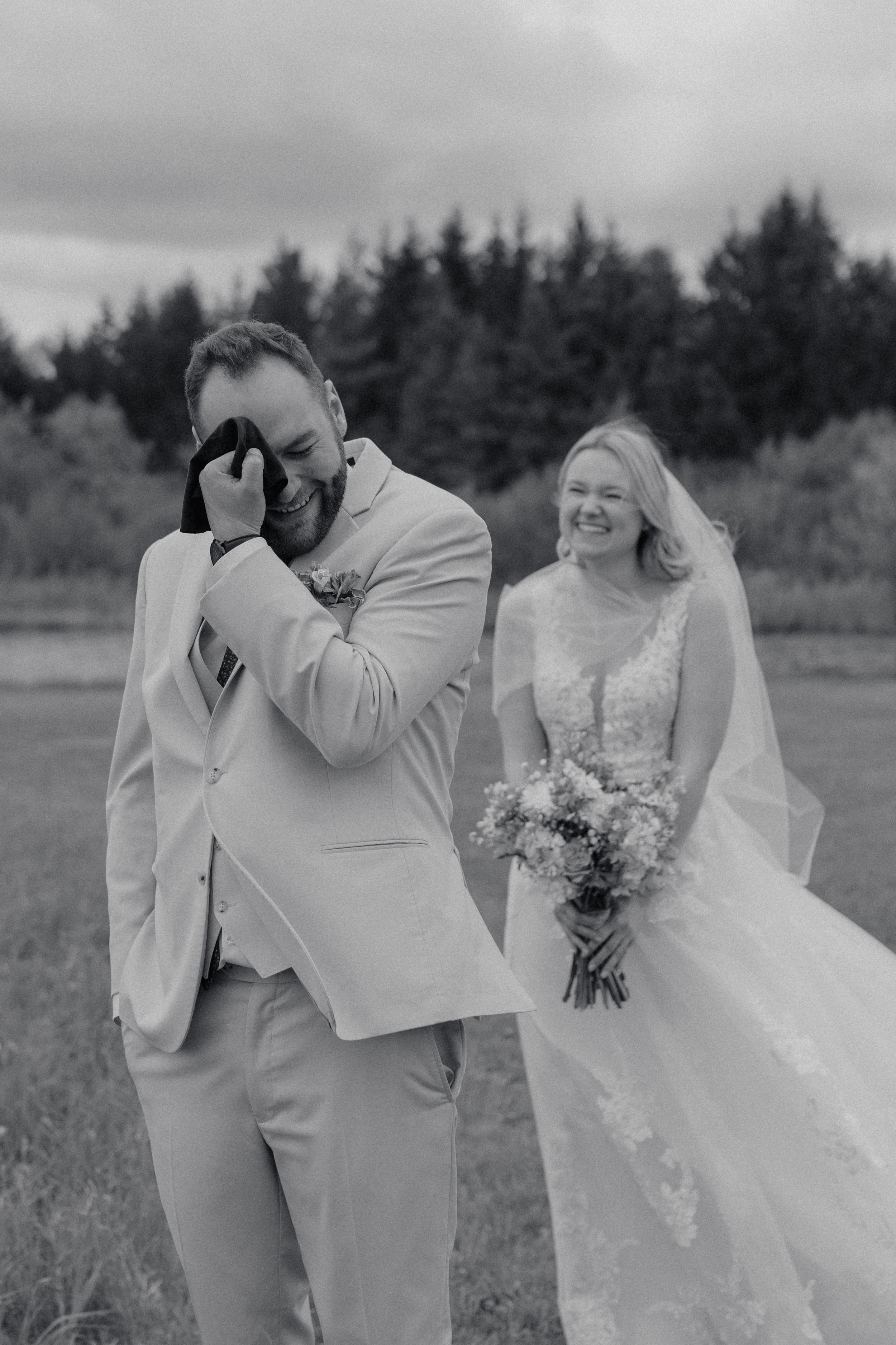 A groom in a light suit is smiling and wiping tears from his eye while a bride in a wedding dress and veil stands behind him, holding a bouquet and smiling in a field with trees in the background.