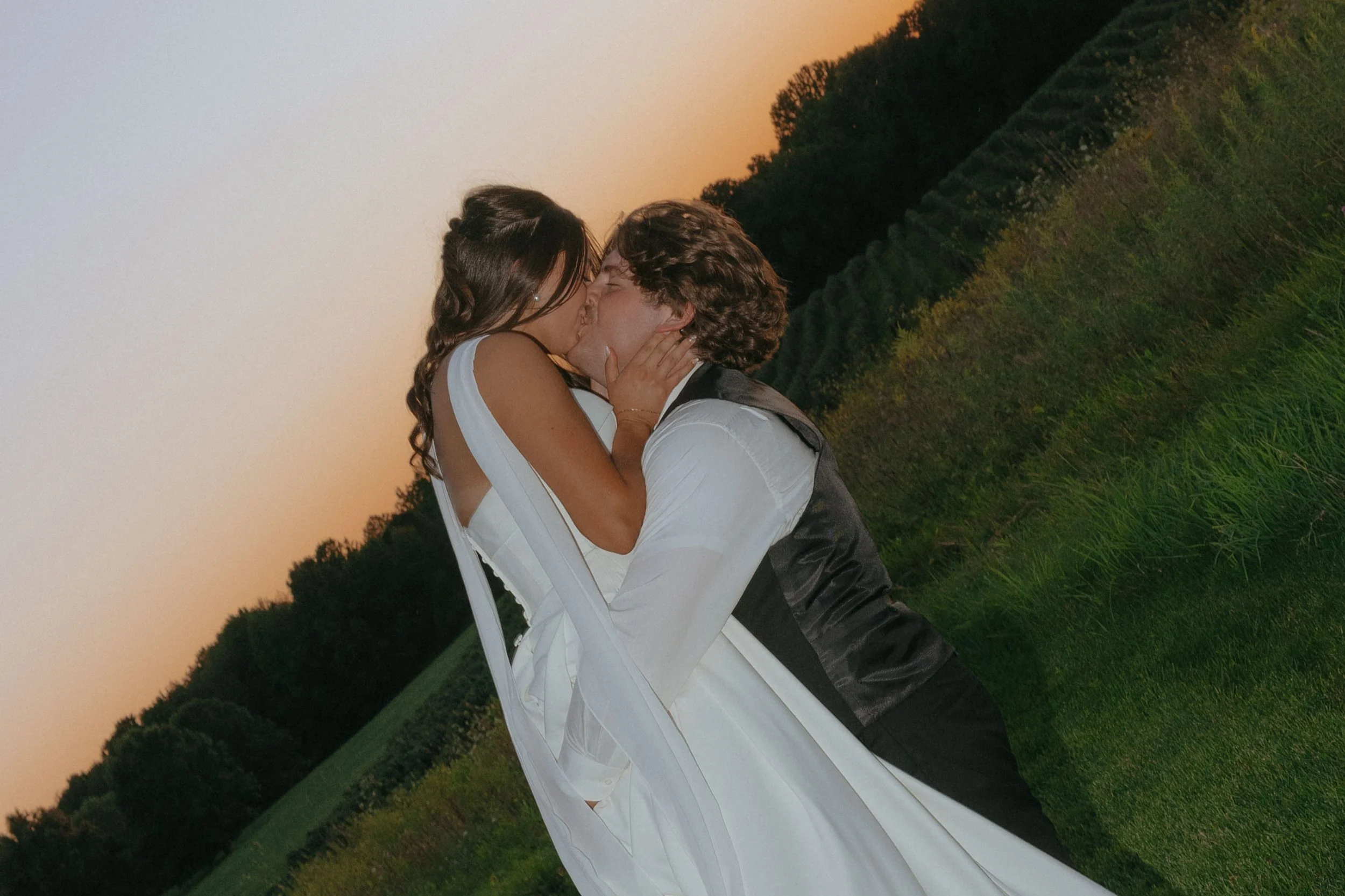 A couple in wedding attire sharing a kiss outdoors during sunset with green trees and fields in the background.