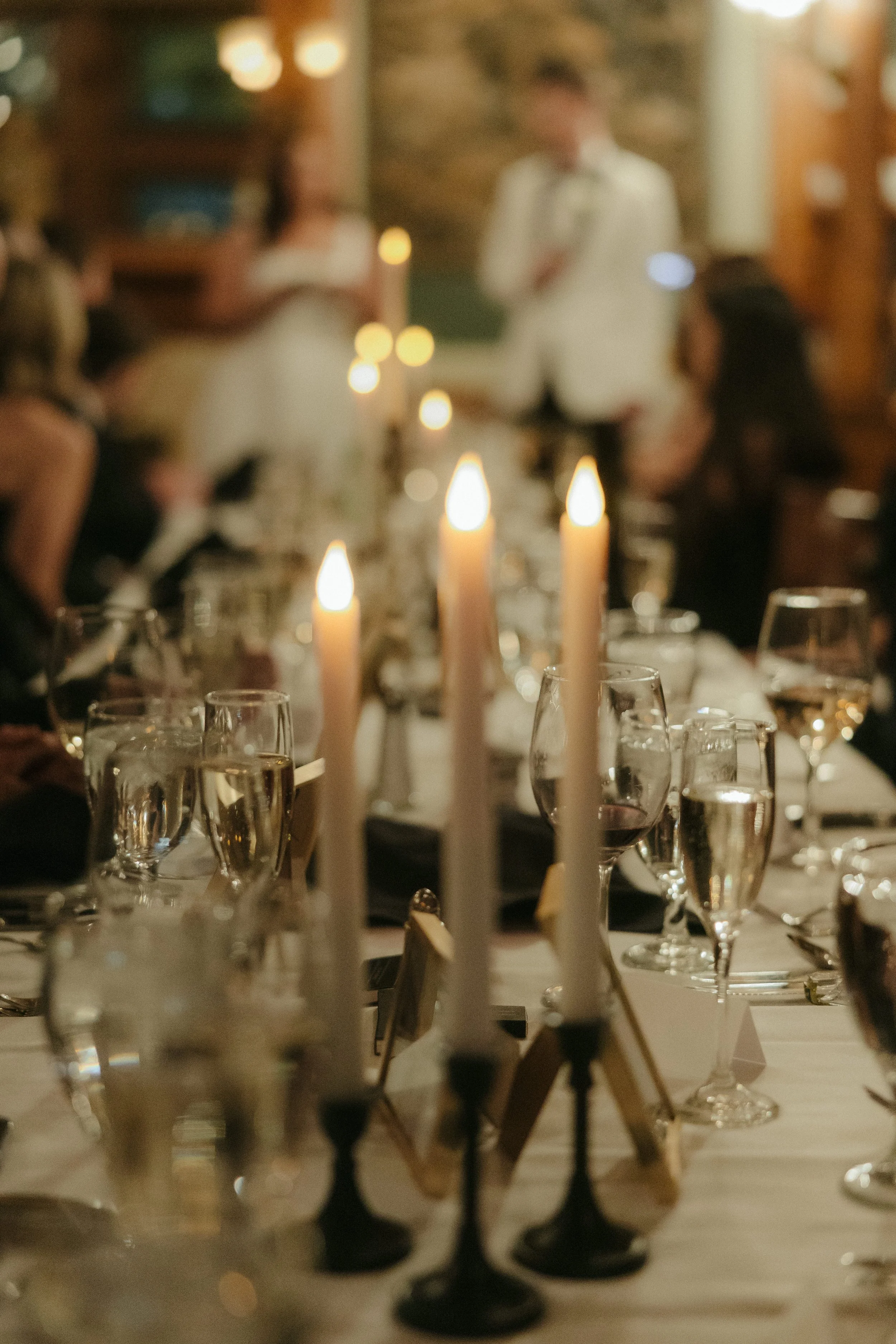 A formal dinner setup with candles, wine glasses, and people in the background at a gathering or celebration.