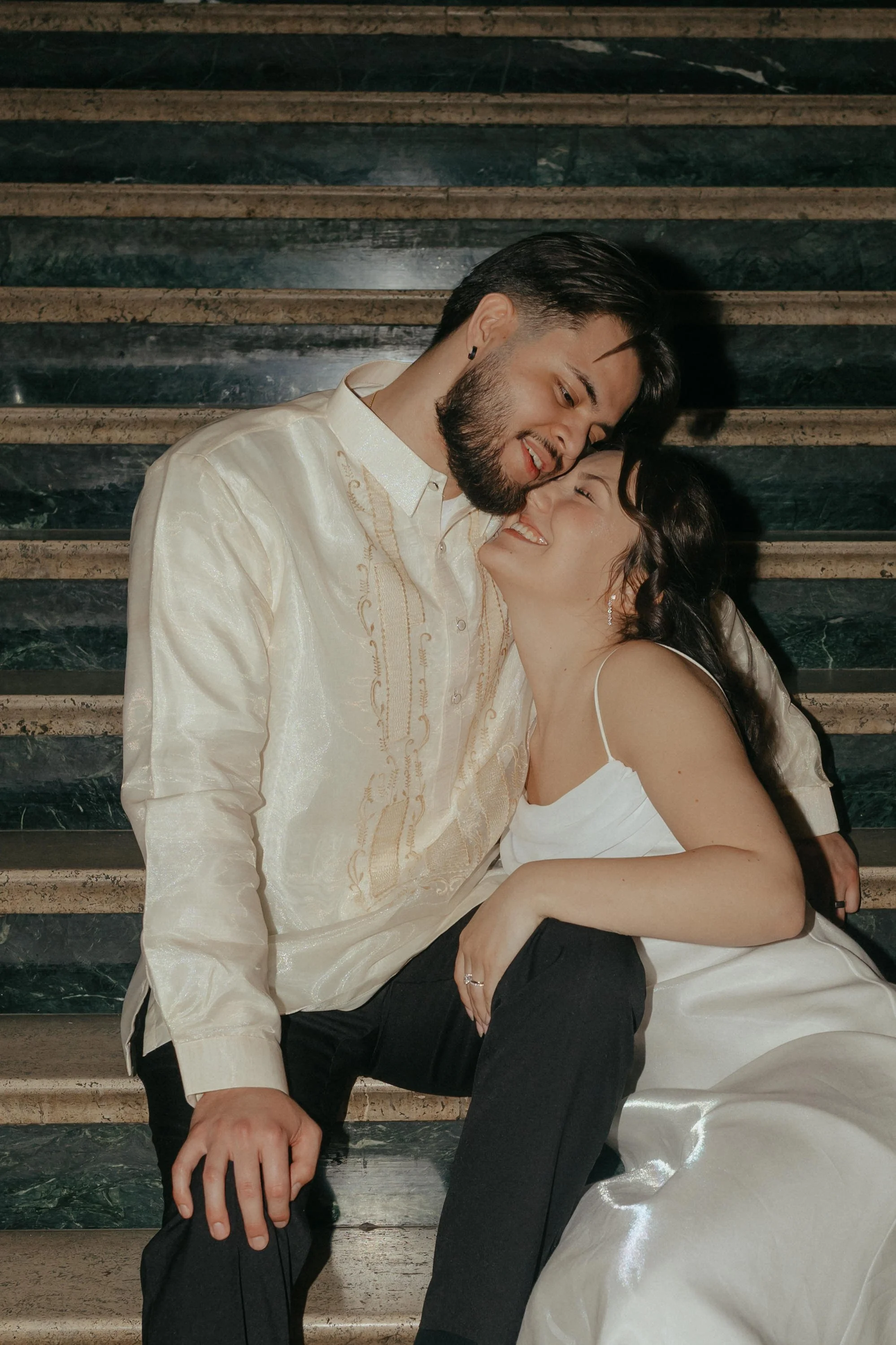 A couple sitting on stairs, smiling and leaning their foreheads together, dressed in elegant clothing.