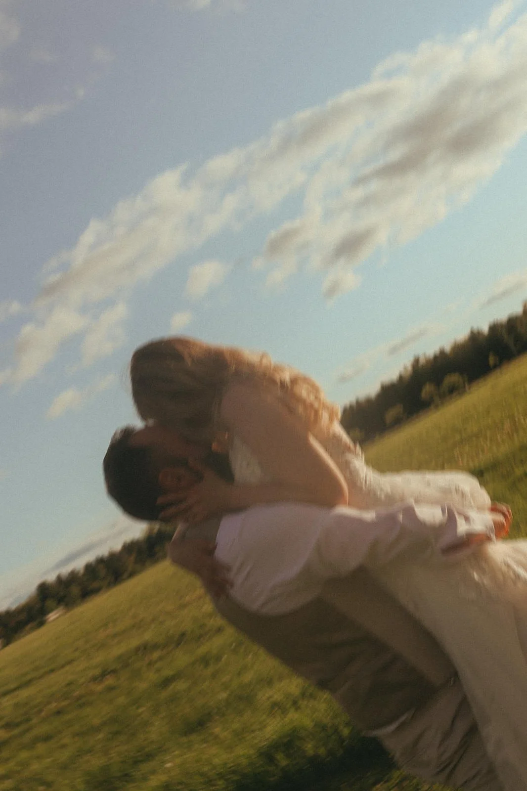A couple kissing outdoors in a grassy field with a blue sky and white clouds overhead.