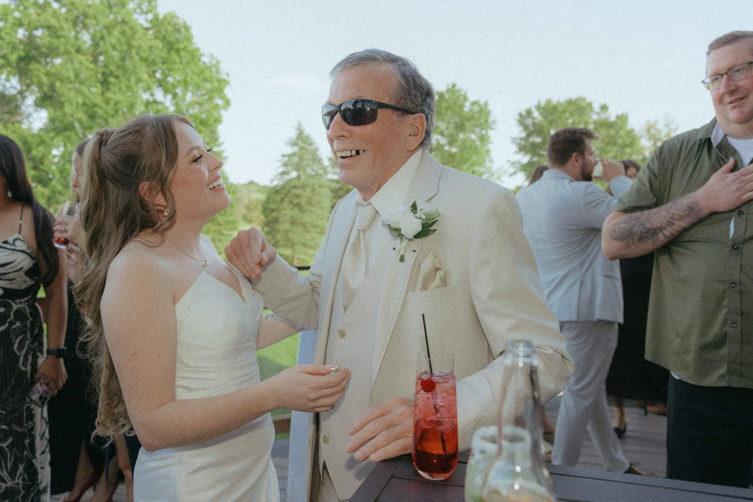 A bride and an elderly man in a white suit are dancing and smiling at an outdoor wedding reception. The bride has long, wavy hair and is wearing a strapless white dress. The elderly man has short gray hair and is wearing sunglasses, a boutonnière, an