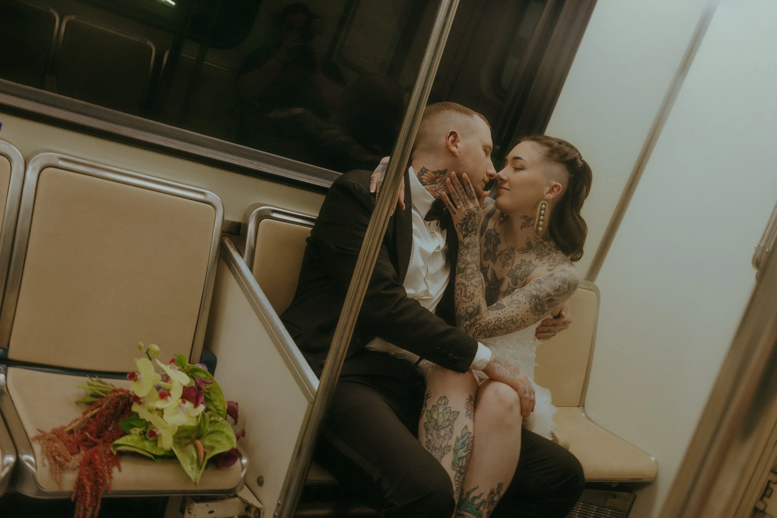 A tattooed couple dressed in formal attire, the man in a black tuxedo and the woman in a white dress, sitting close together on a subway train, looking into each other's eyes, with a bouquet of flowers on the seat beside them.