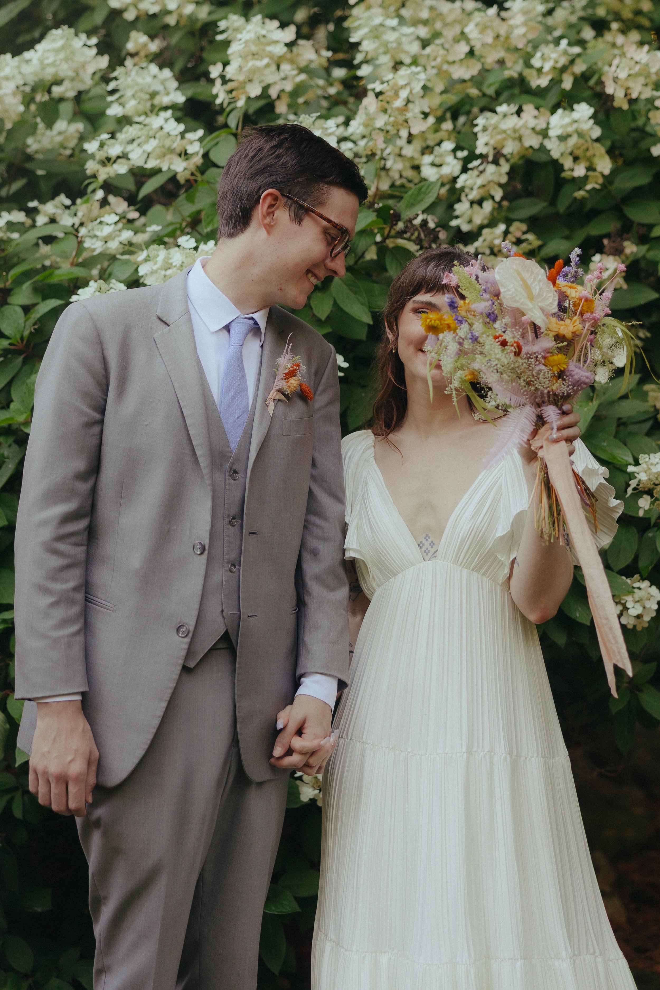 A bride and groom holding hands, standing in front of lush green foliage with white flowers. The bride is holding a colorful bouquet and smiling, while the groom is smiling and looking at her.