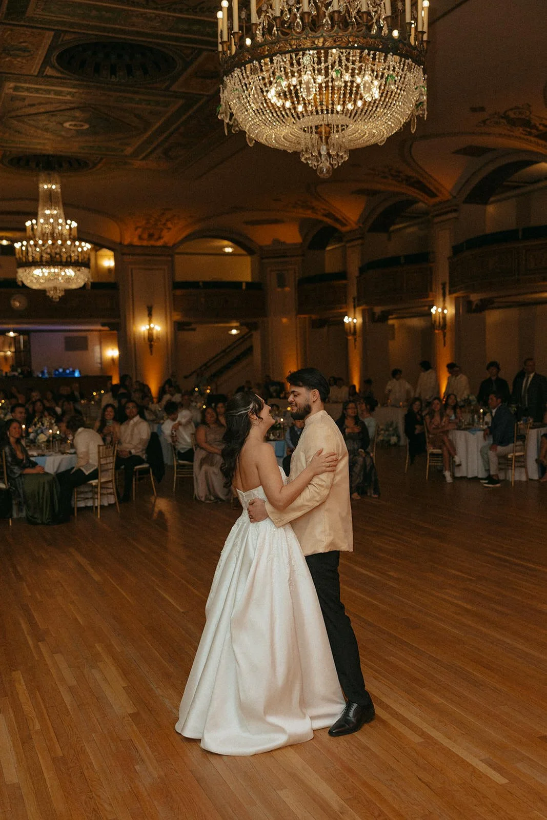 A bride and groom are dancing at their wedding reception in an elegant banquet hall with grand chandeliers and warm lighting. Guests are seated at round tables in the background.