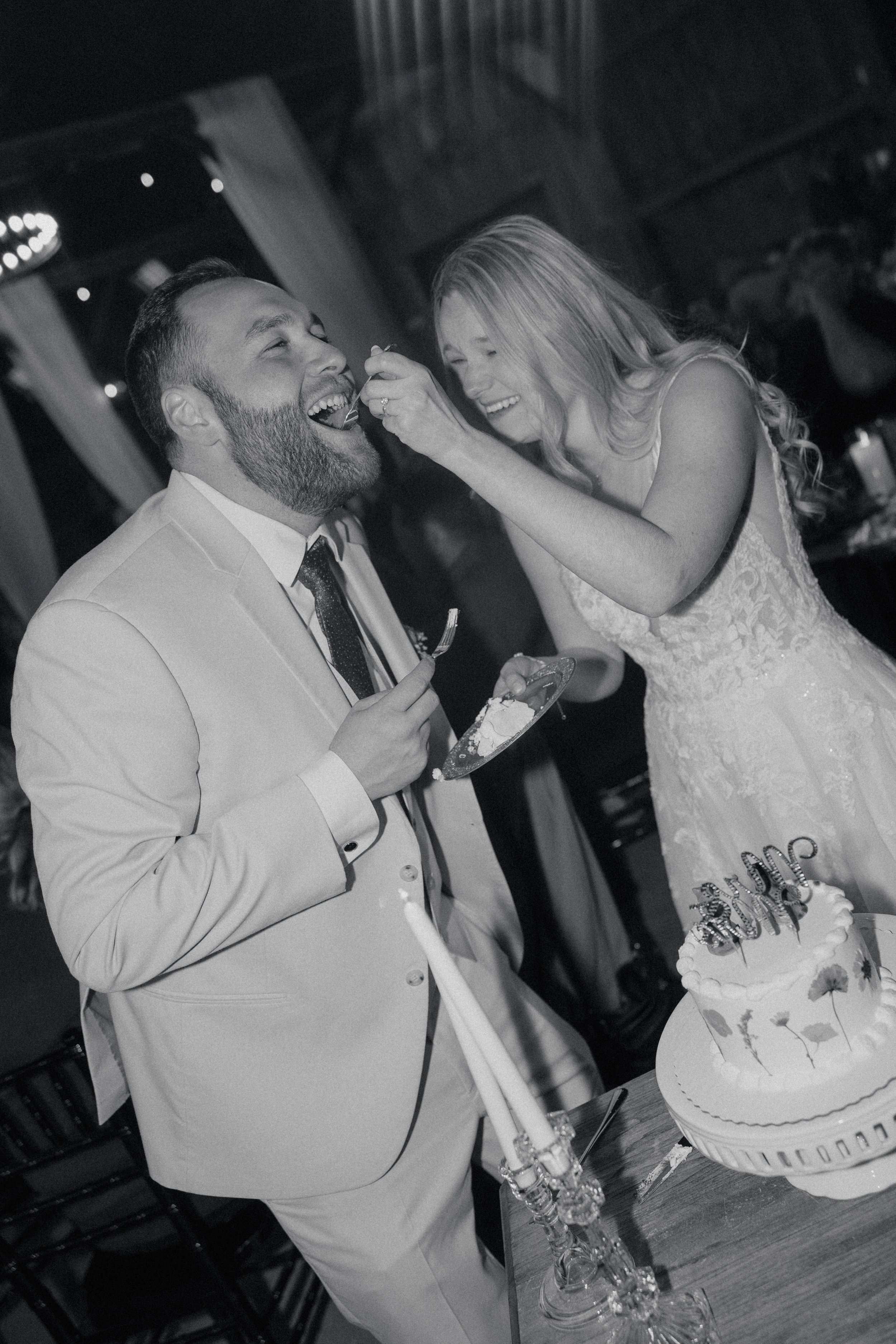 A bride and groom celebrating their wedding with cake, at a reception. The bride is feeding cake to the groom, who is smiling and holding a fork, while standing by a decorated wedding cake.
