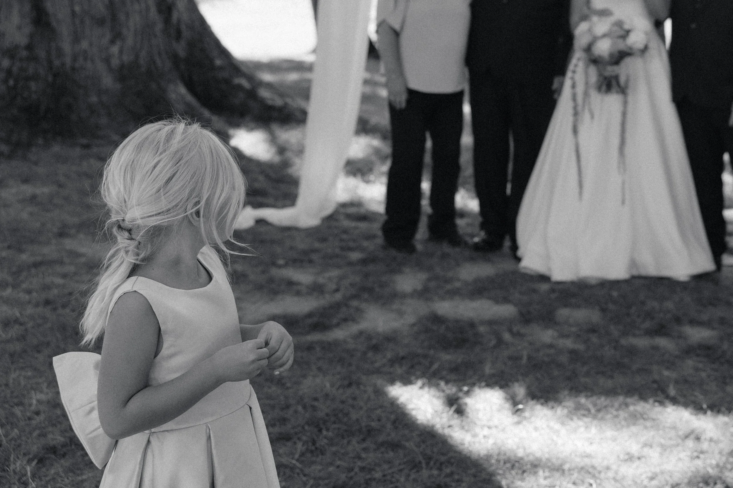 A young girl with blonde hair in a braid, wearing a sleeveless dress, stands outdoors at a wedding ceremony, looking towards the couple getting married.