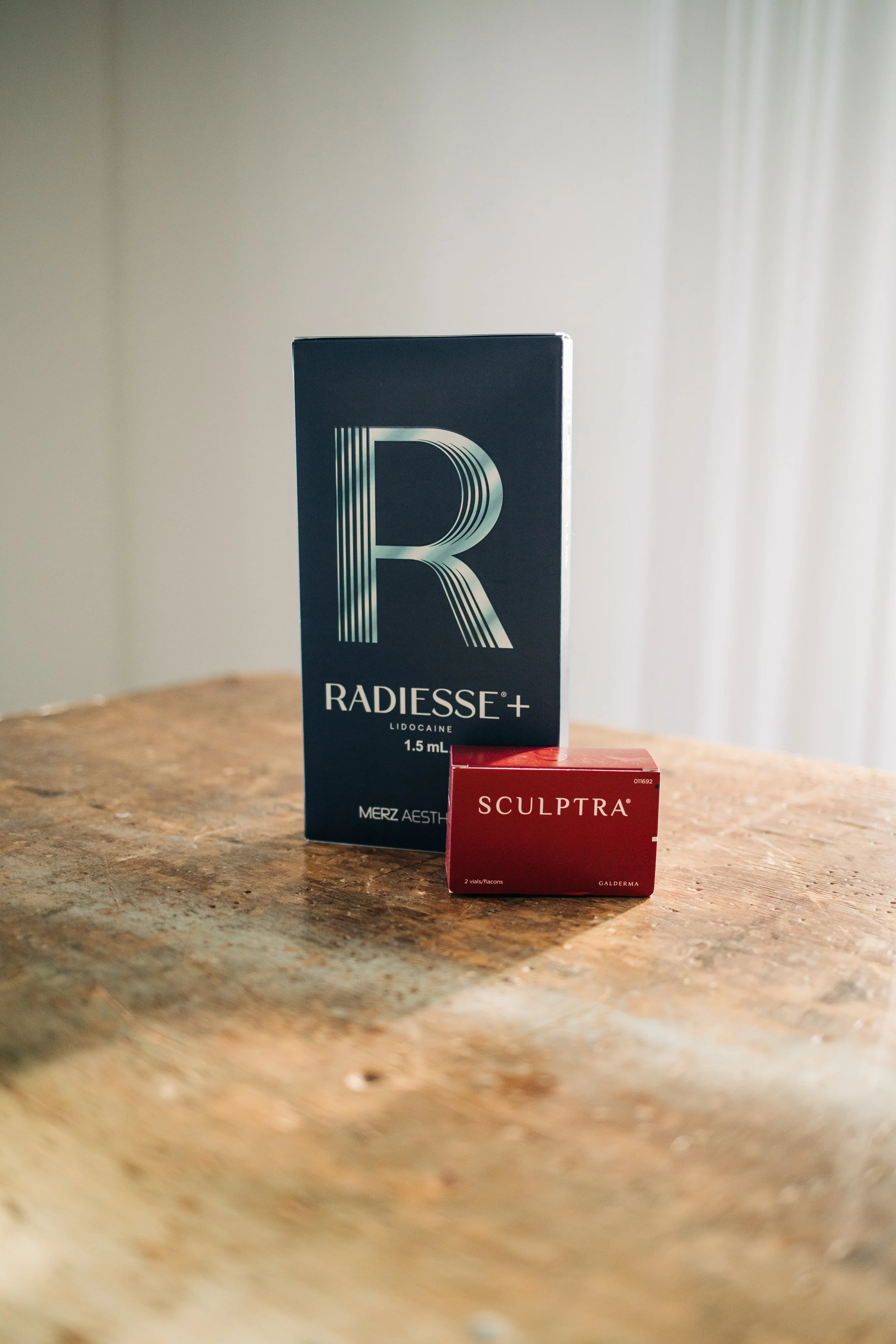 A box of Radiesse+ dermal filler and a box of Sculptra cosmetic injections placed on a wooden surface with a plain background.