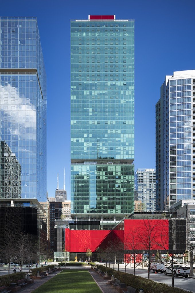 A tall modern glass skyscraper with a red rooftop section, surrounded by similar glass buildings in an urban cityscape, with a landscaped park in the foreground.