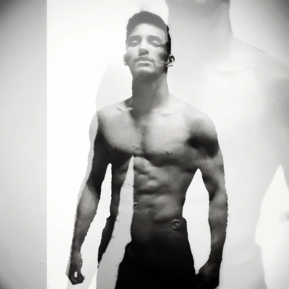 Black and white photo of a shirtless young man with defined abs and chest, standing against a plain background.