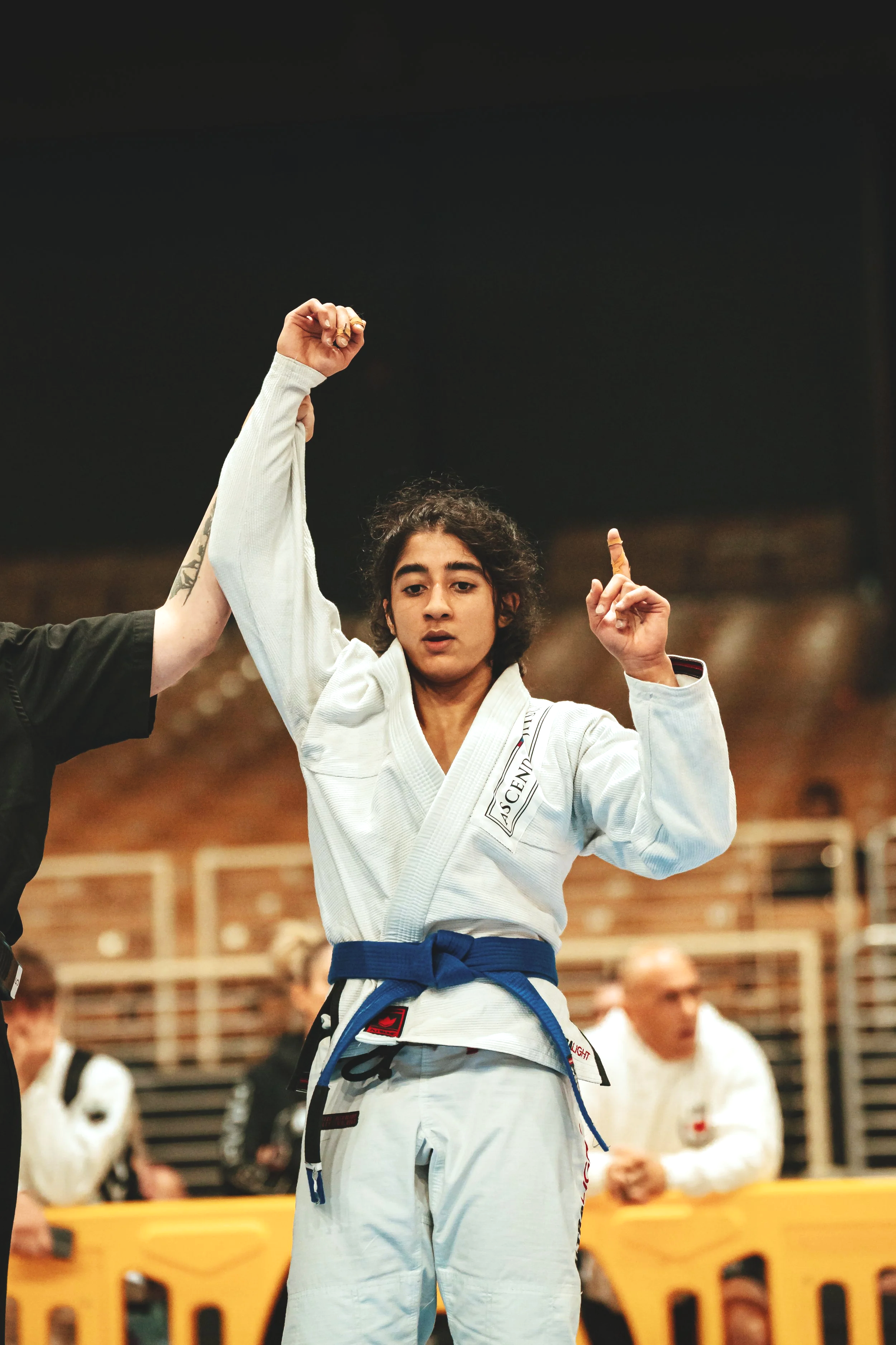 Ascend Jiu-Jitsu's Coach Ananya in a white gi with a blue belt raising her fist in a victory pose during a tournament.