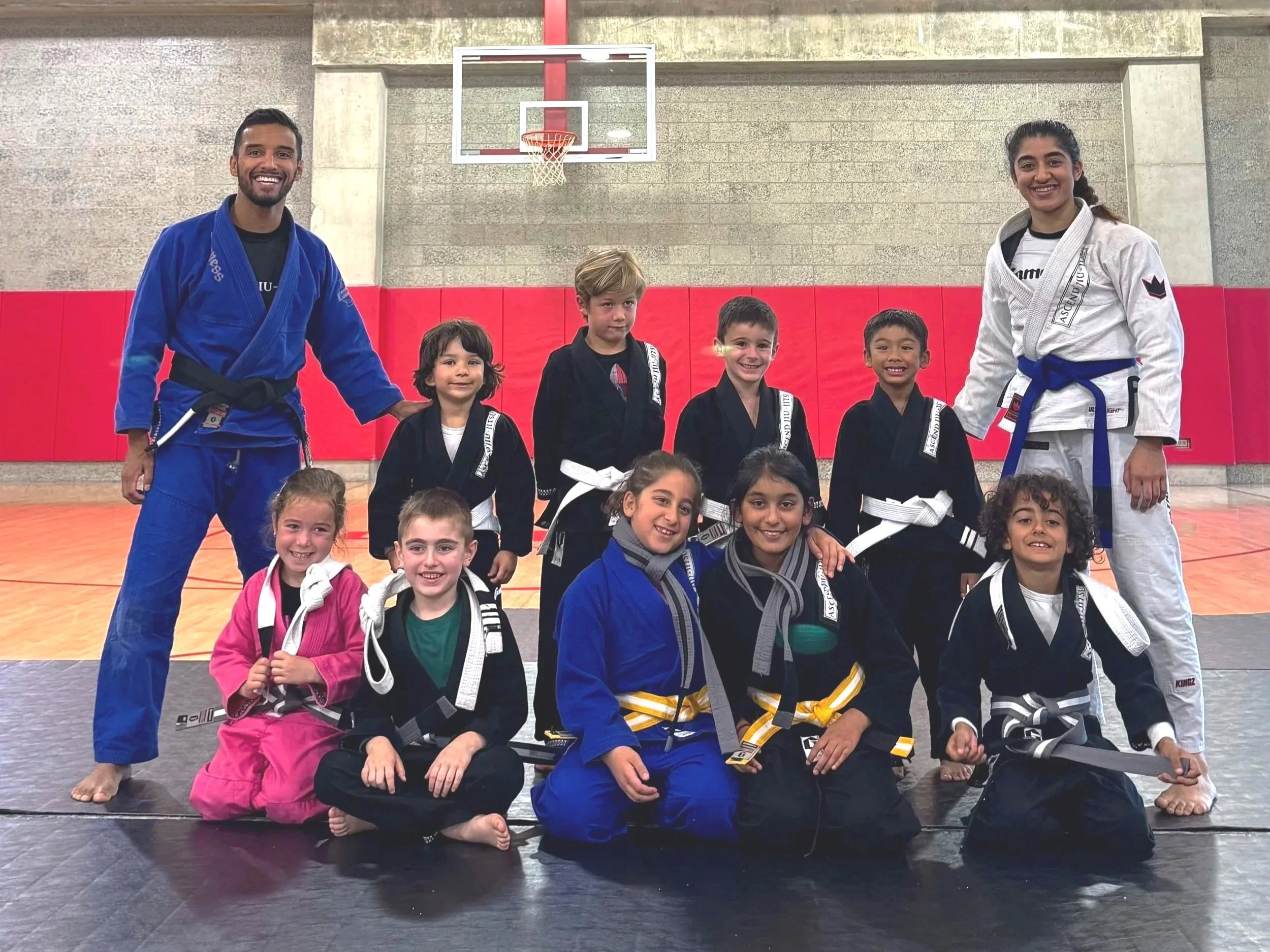 Group photo of children and two adult martial arts instructors in a gym, with children dressed in martial arts uniforms and an orange and a yellow belt. The instructors are wearing blue and white gis, respectively. Ascend Jiu-Jitsu team photo