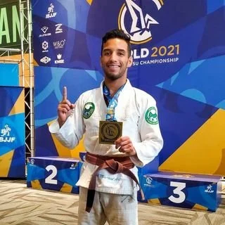 A male jiu-jitsu athlete holding a gold medal and a trophy, standing on a podium at a 2021 BJJ championship event.
