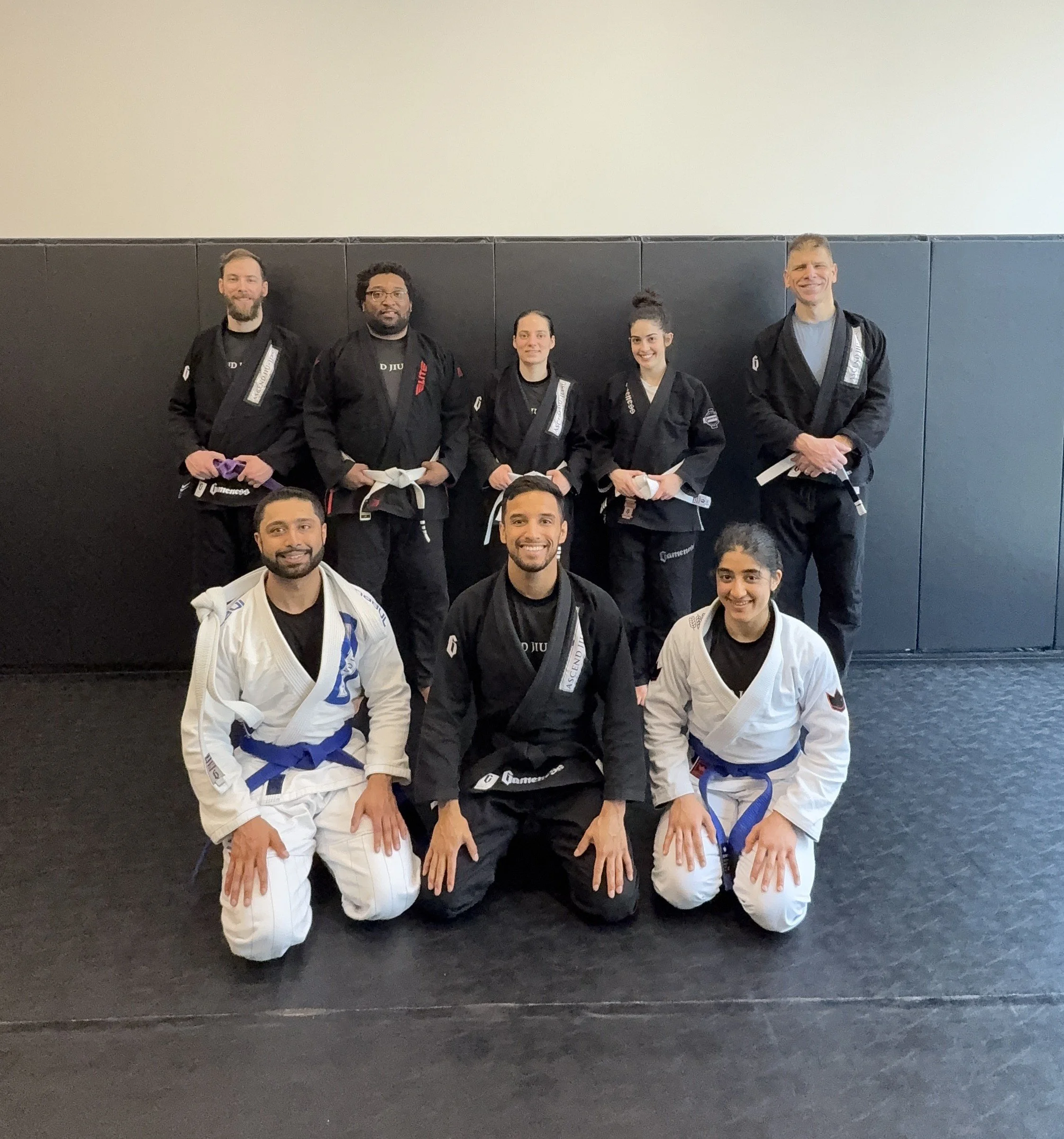 Ascend Jiu-Jitsu group picture of eight people, five men and three women, wearing martial arts uniforms with belts, posing in a gym or dojo with black mats and a black wall in the background. 