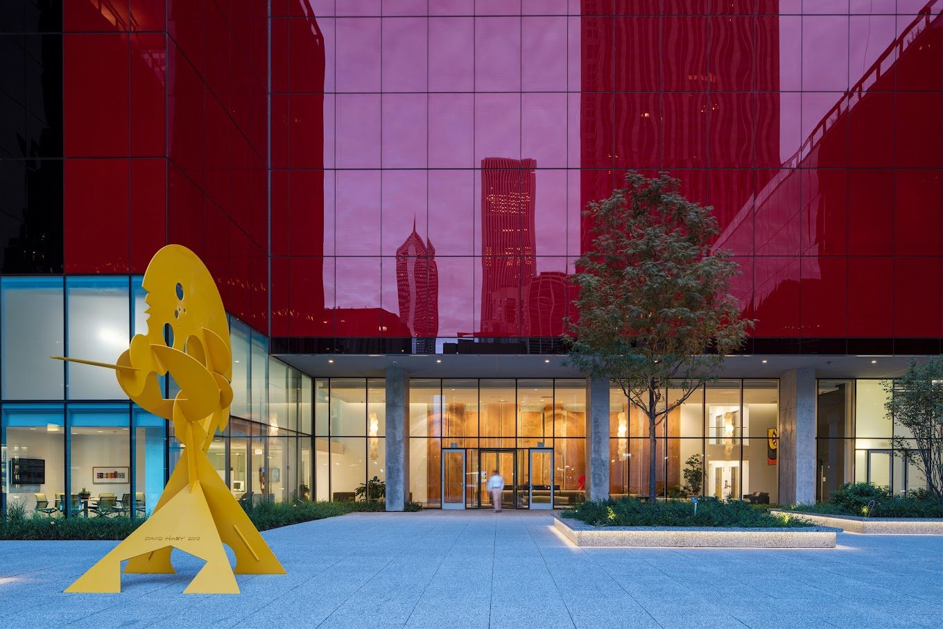 A modern city building with a red reflective glass facade, displaying the downtown skyline and the Freedom Tower in the background. In the foreground, there is a bright yellow abstract sculpture of a person playing a guitar outside the entrance.