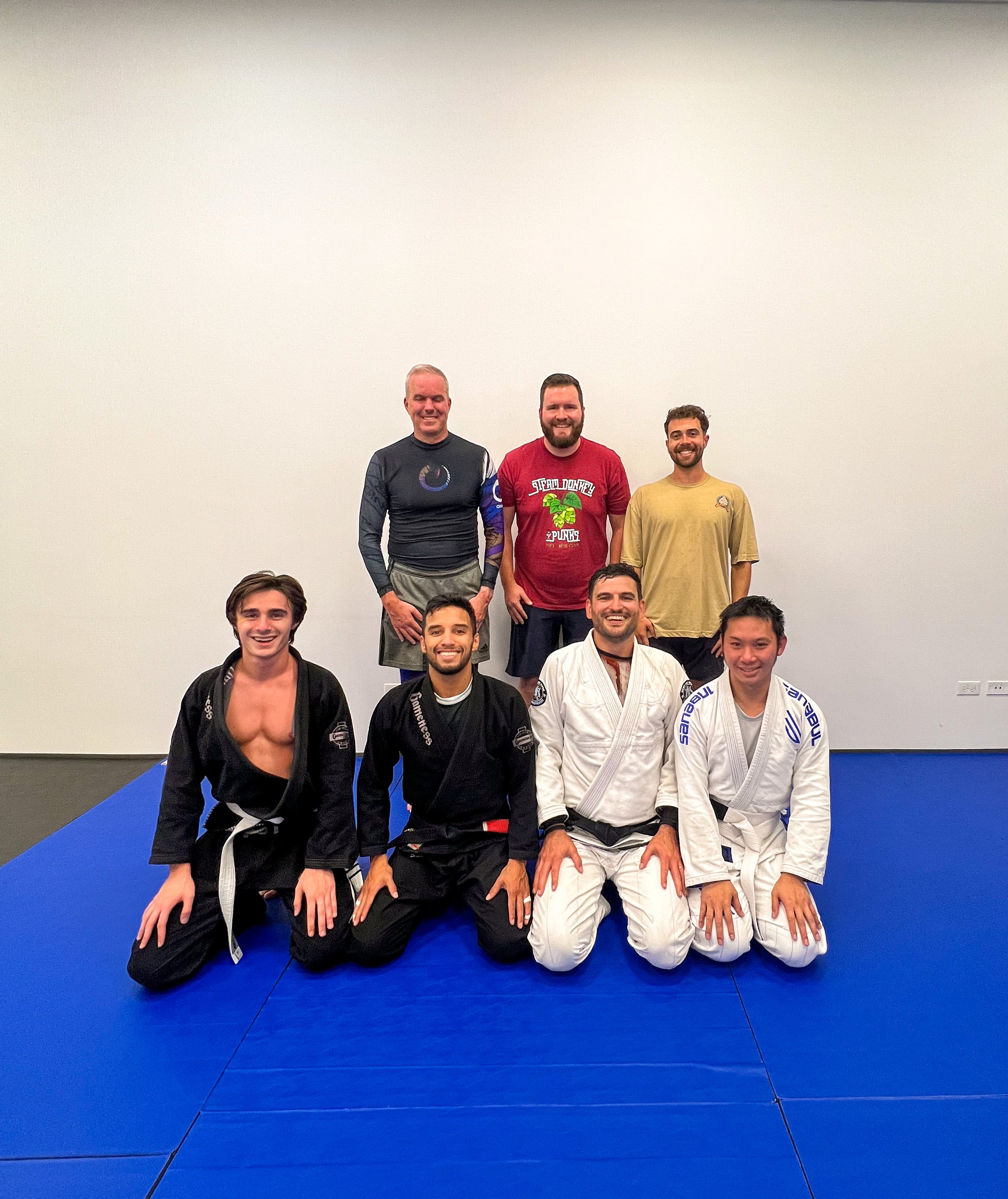 Seven people, dressed in gi and casual clothing, posing for a group photo on a martial arts mat in a gym with plain white walls.