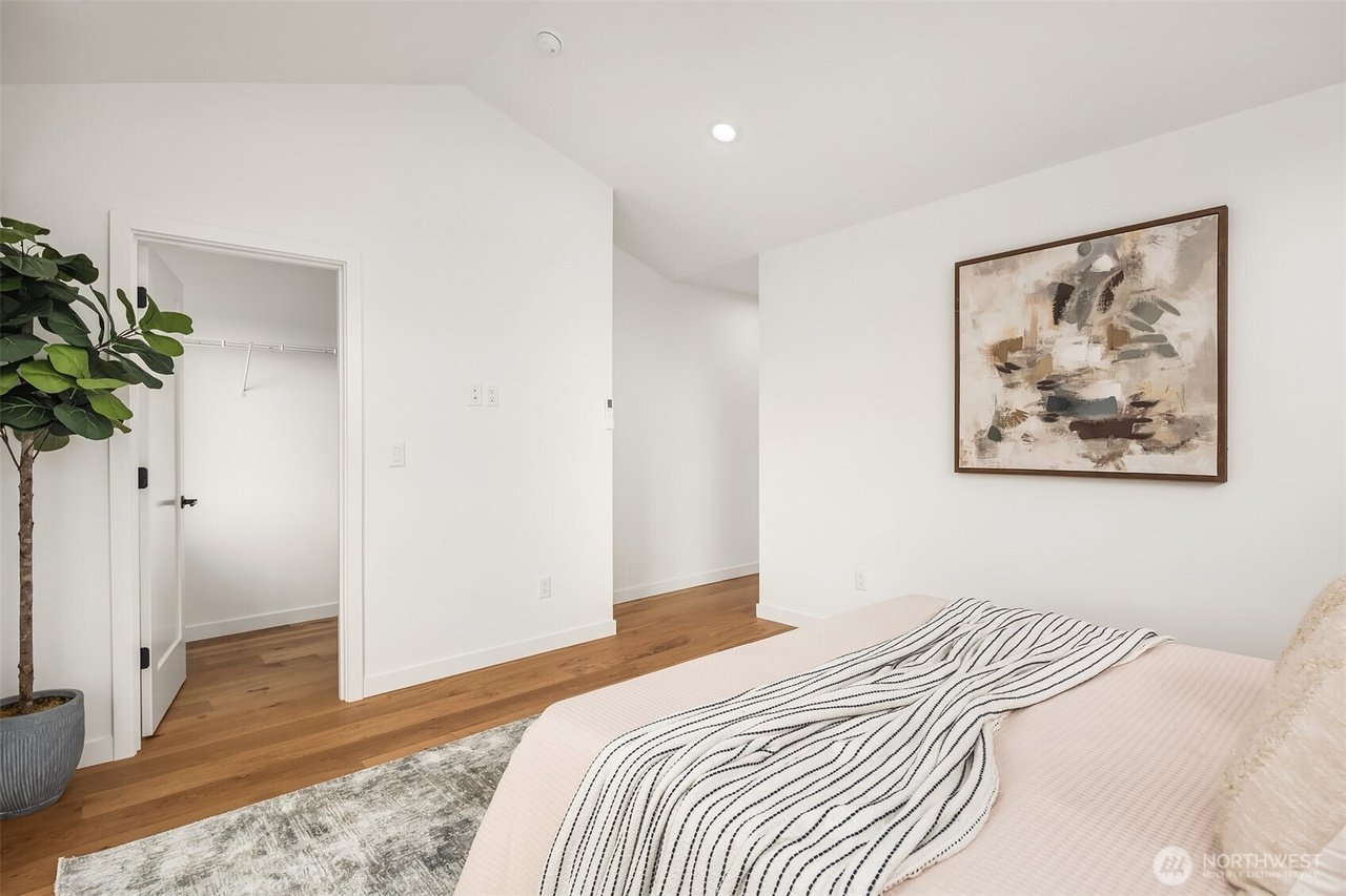 Modern bedroom with white walls, hardwood floors, abstract artwork, and a potted plant. The room has an open closet door and a neatly made bed with a striped throw blanket.