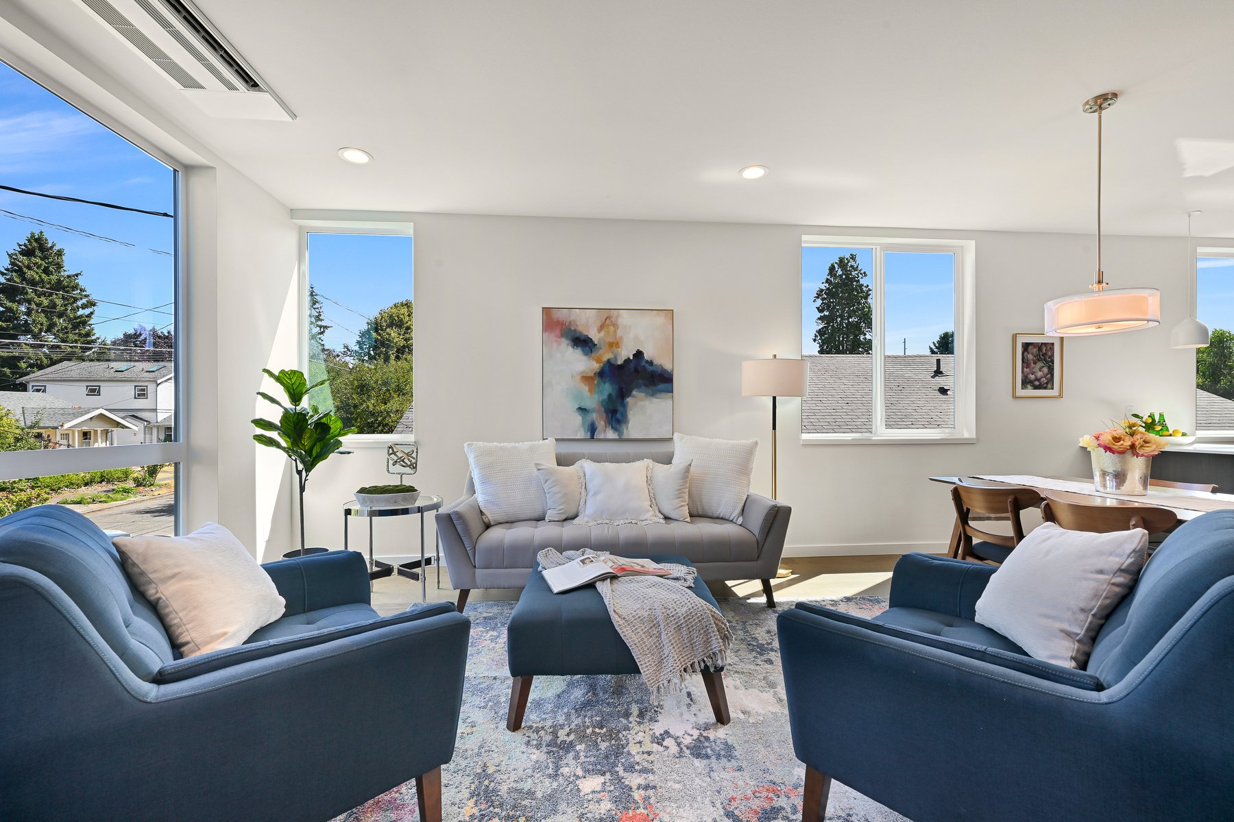 Modern living room with blue armchairs, gray sofa, abstract art, and large windows showing outdoor view. A dining table with chairs and pendant light is visible in the background, along with a decorative plant and a floor lamp.