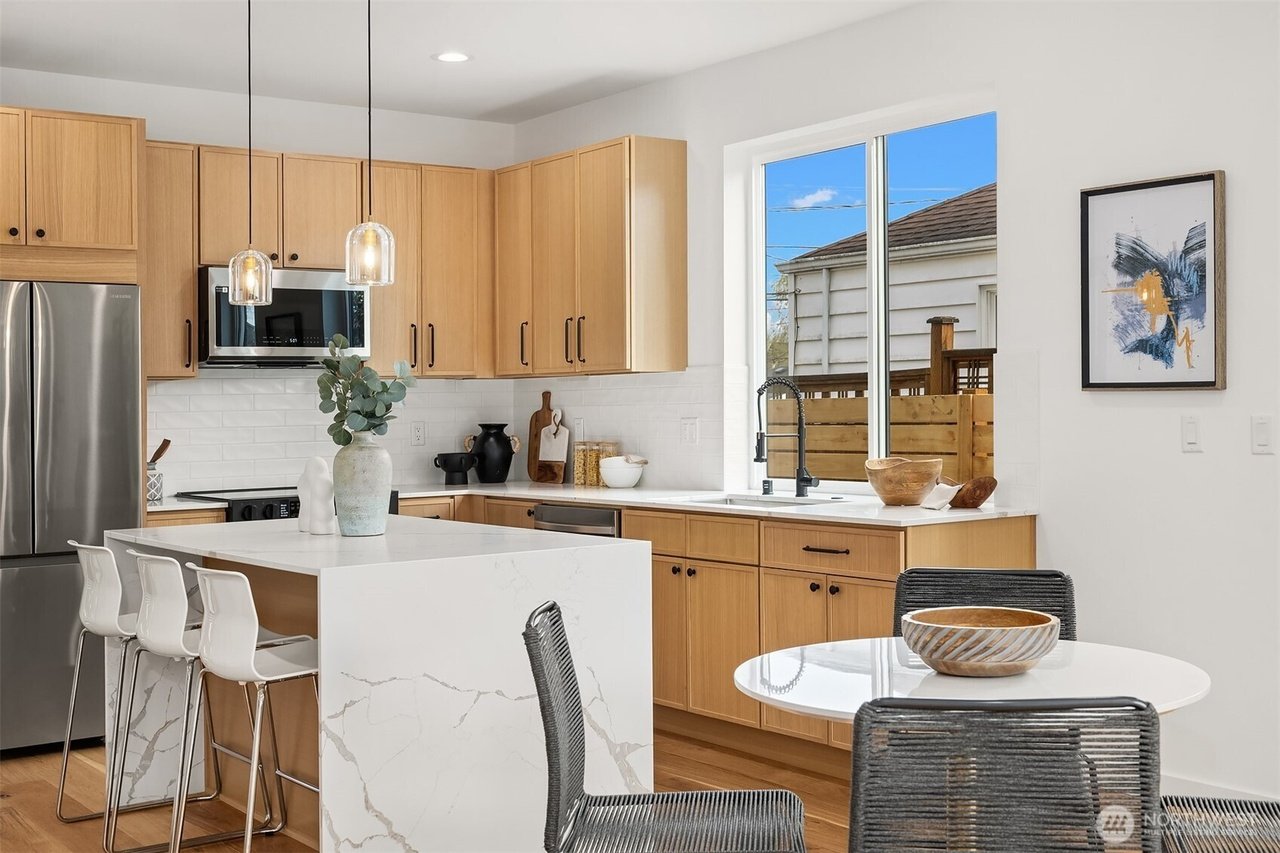 Modern kitchen with light wood cabinets, white quartz counters, stainless steel appliances, and a large island with bar stools. Pendant lights hang above the island, and a round dining table with wicker chairs is in the foreground. A decorative plant