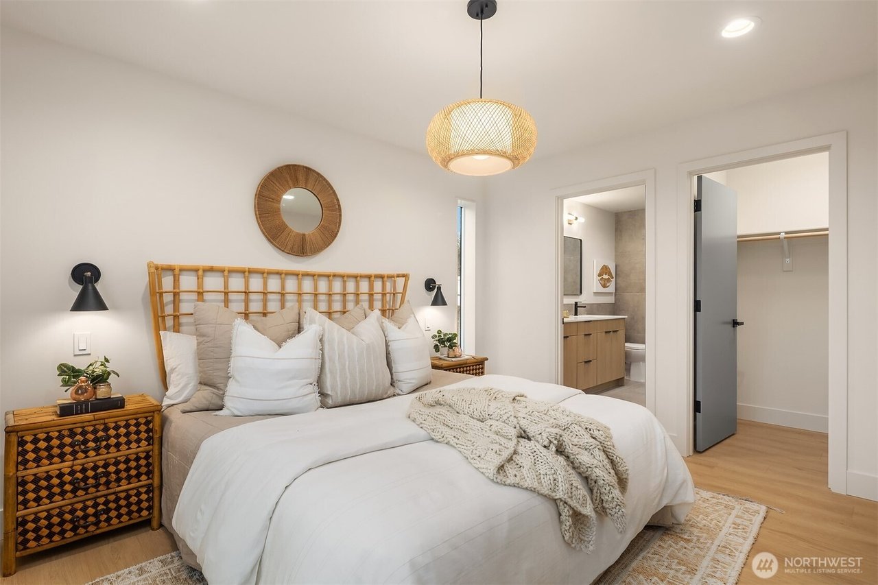 Modern bedroom with wicker headboard and white bedding, accented by a knit throw. Nightstand with decorative plants and books. Circular wall mirror above bed. Adjacent view into a bathroom and walk-in closet. Overhead woven light fixture.