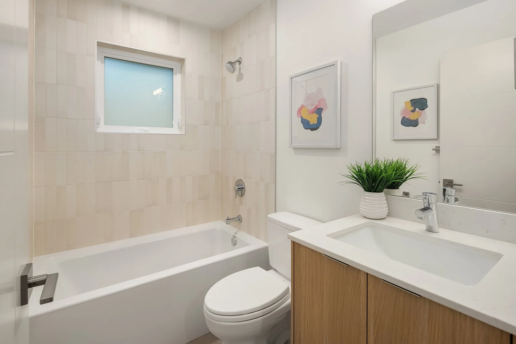 Modern bathroom with beige tiled shower area, white toilet, wooden vanity with white sink, large mirror, green plant, and framed artwork on white walls.
