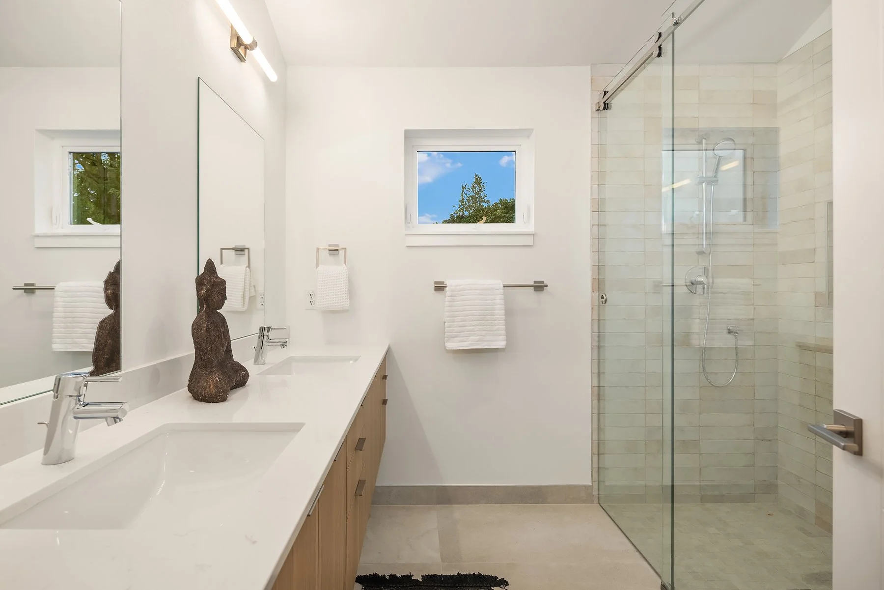 Modern bathroom with dual sinks, wooden vanity, decorative statues, towel rack, window showing blue sky, and glass-enclosed shower with beige tiles.