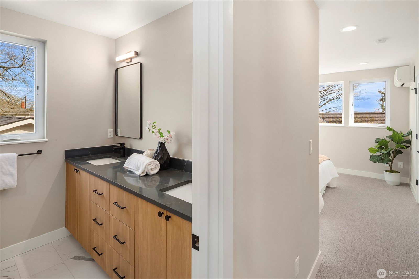 Modern bathroom and bedroom view; bathroom features a wooden vanity with dual sinks, black countertop, and a rectangular mirror; white towel and vase with flowers on counter; bedroom visible through doorway, with light walls, carpet, and large window